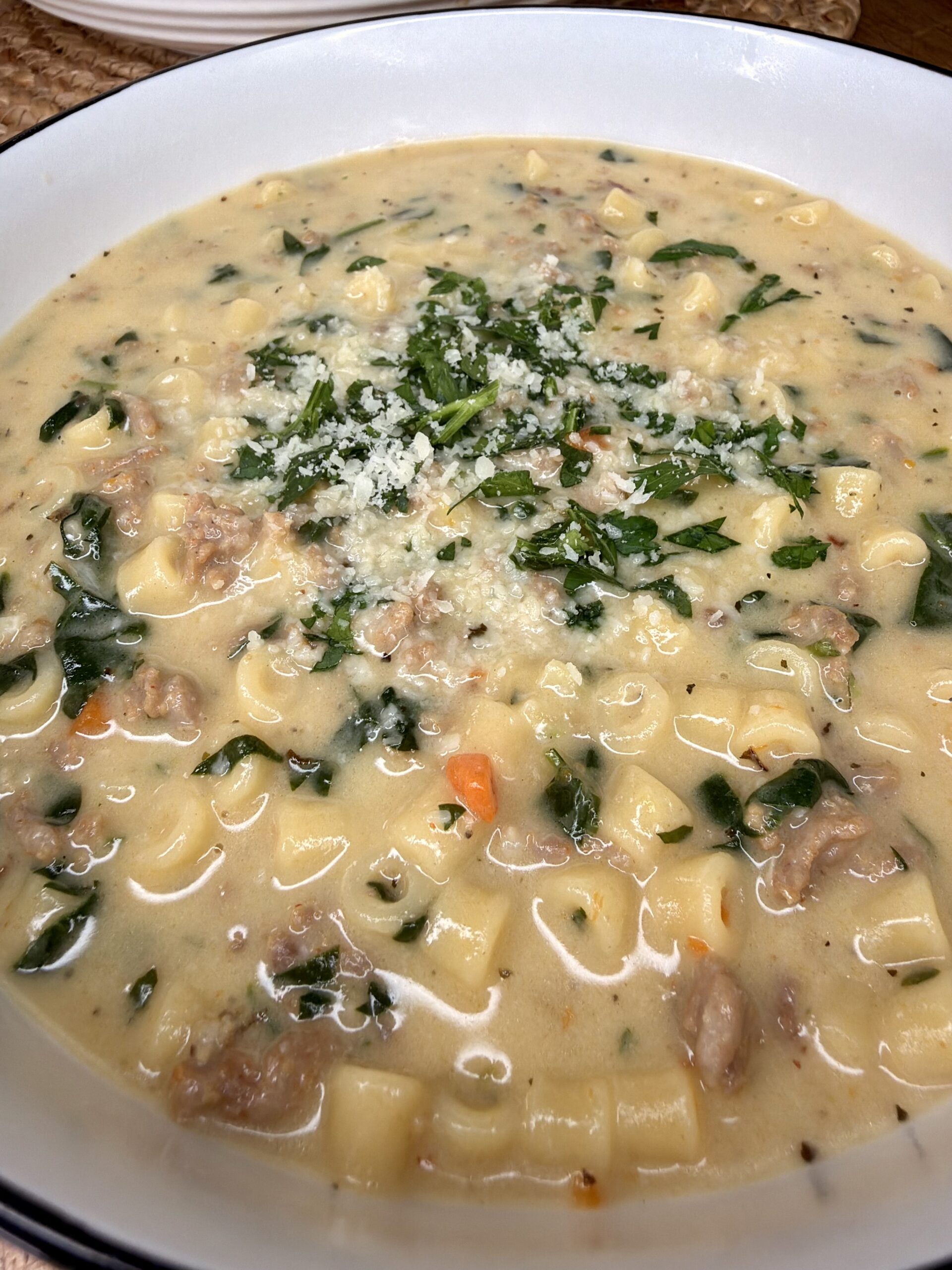 A creamy soup with small pasta, ground meat, spinach, and carrots, topped with grated cheese and fresh herbs, served in a white bowl.