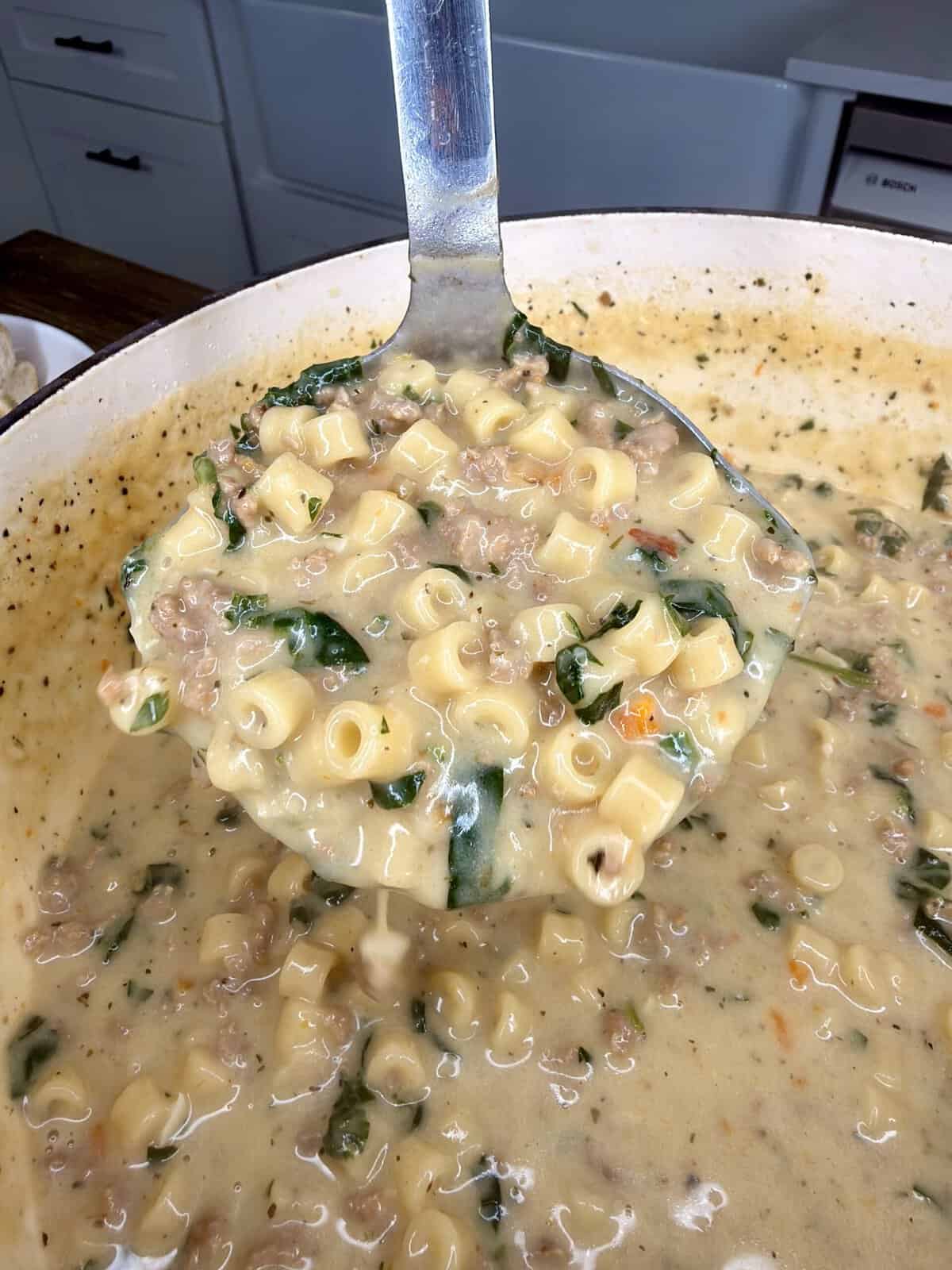 A ladle lifts creamy soup with ditalini pasta, ground meat, spinach, and vegetables from a large pot in a kitchen setting.