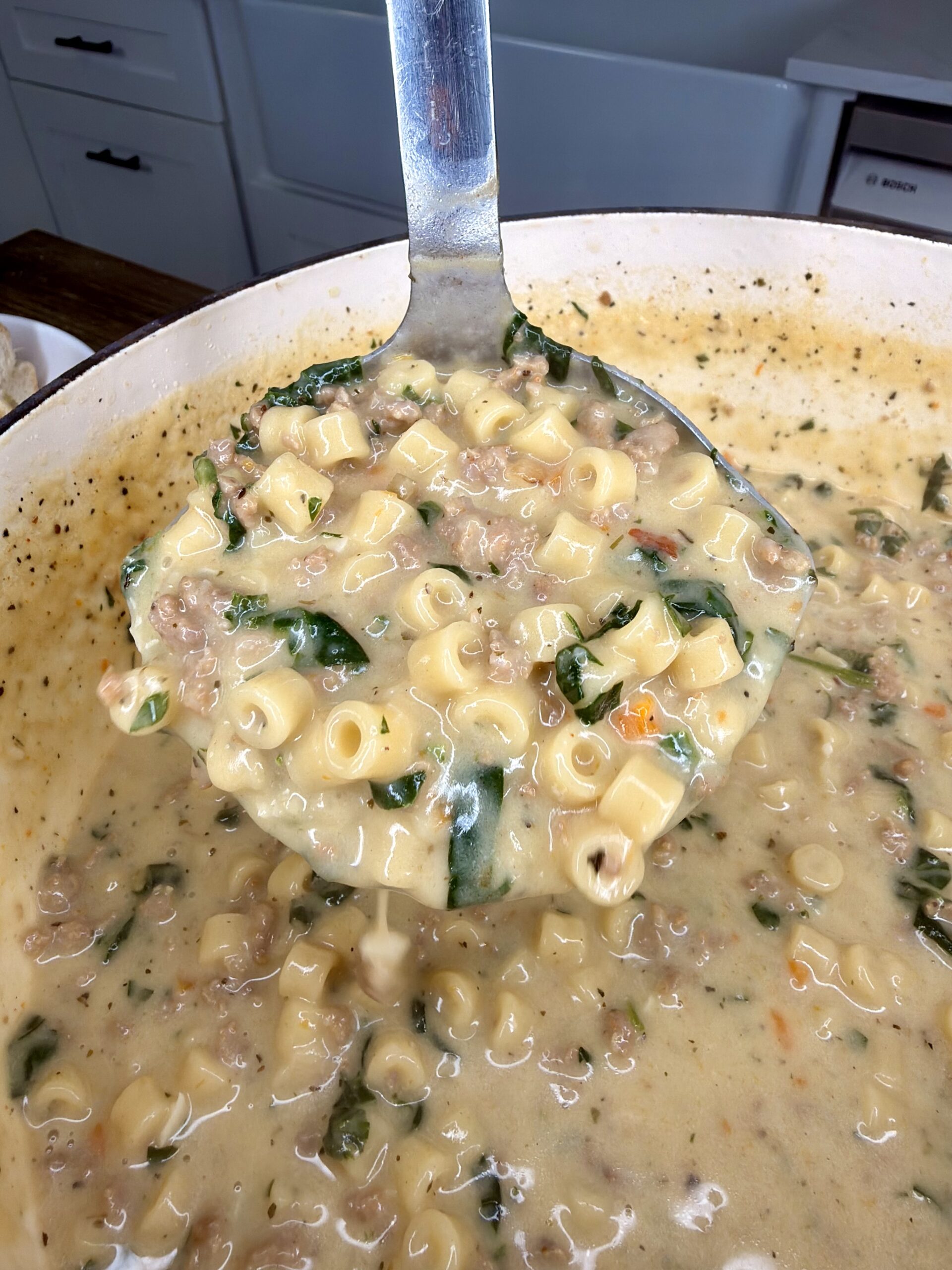 A ladle lifting creamy soup with ditalini pasta, ground meat, chopped spinach, and herbs from a large white pot in a kitchen setting.