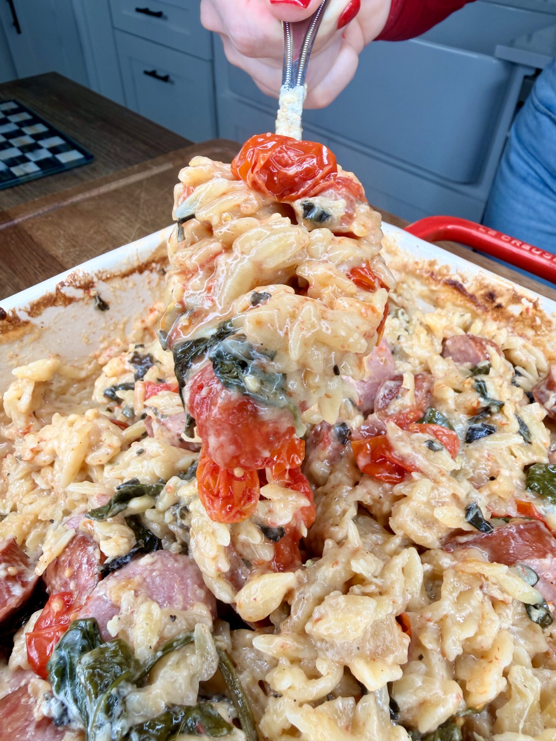 A close-up of a creamy baked pasta dish with spiral noodles, roasted cherry tomatoes, spinach, and sausage being scooped from a red baking dish with a serving utensil.