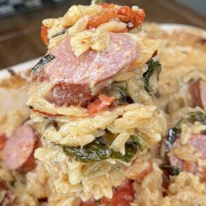 A close-up of a spoonful of cheesy rice casserole with sausage slices, wilted greens, and sun-dried tomatoes being lifted from a baking dish, reminiscent of a Creamy Spinach Artichoke Sausage Orzo Bake.