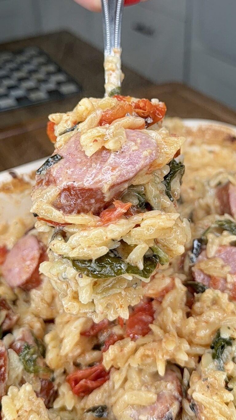 A close-up of a spoonful of cheesy rice casserole with sausage slices, wilted greens, and sun-dried tomatoes being lifted from a baking dish, reminiscent of a Creamy Spinach Artichoke Sausage Orzo Bake.