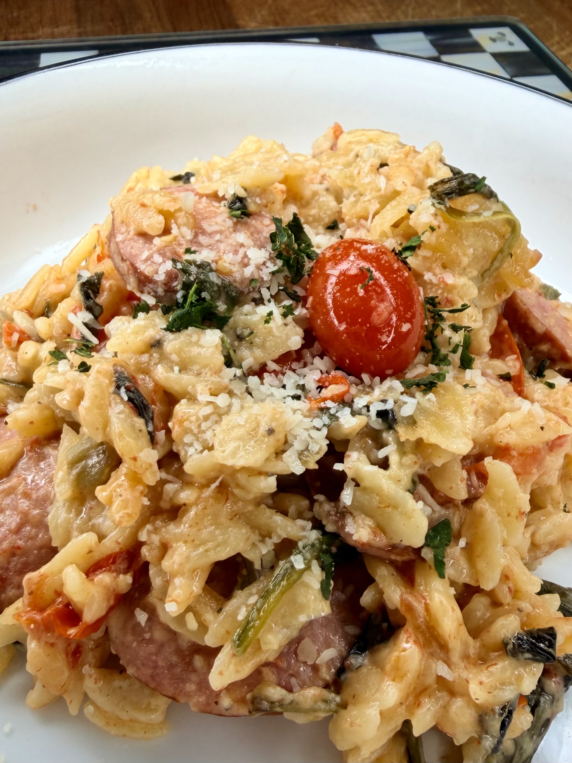 A close-up of a creamy pasta dish with bow-tie pasta, sliced sausage, cherry tomatoes, wilted greens, and grated cheese, served on a white plate.