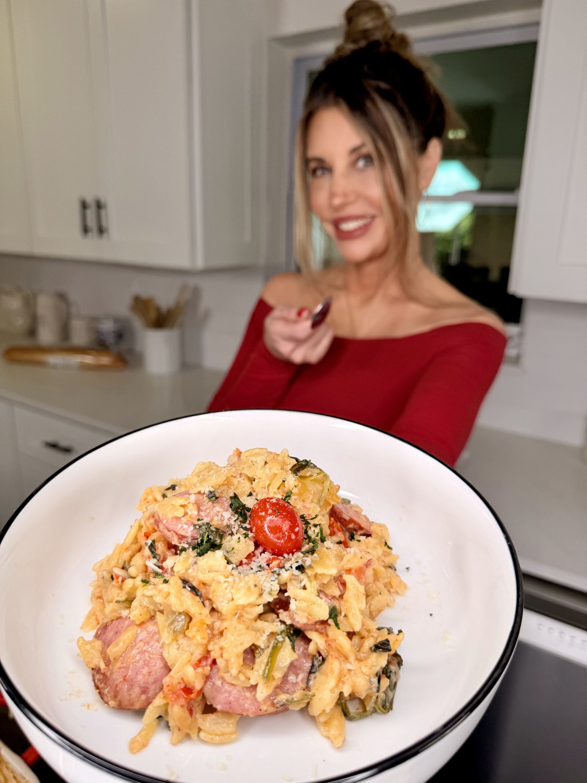 A woman in a red top stands in a modern kitchen, smiling and holding a fork. In the foreground, a close-up of a bowl of creamy pasta with vegetables, sausage, and cherry tomatoes is shown.