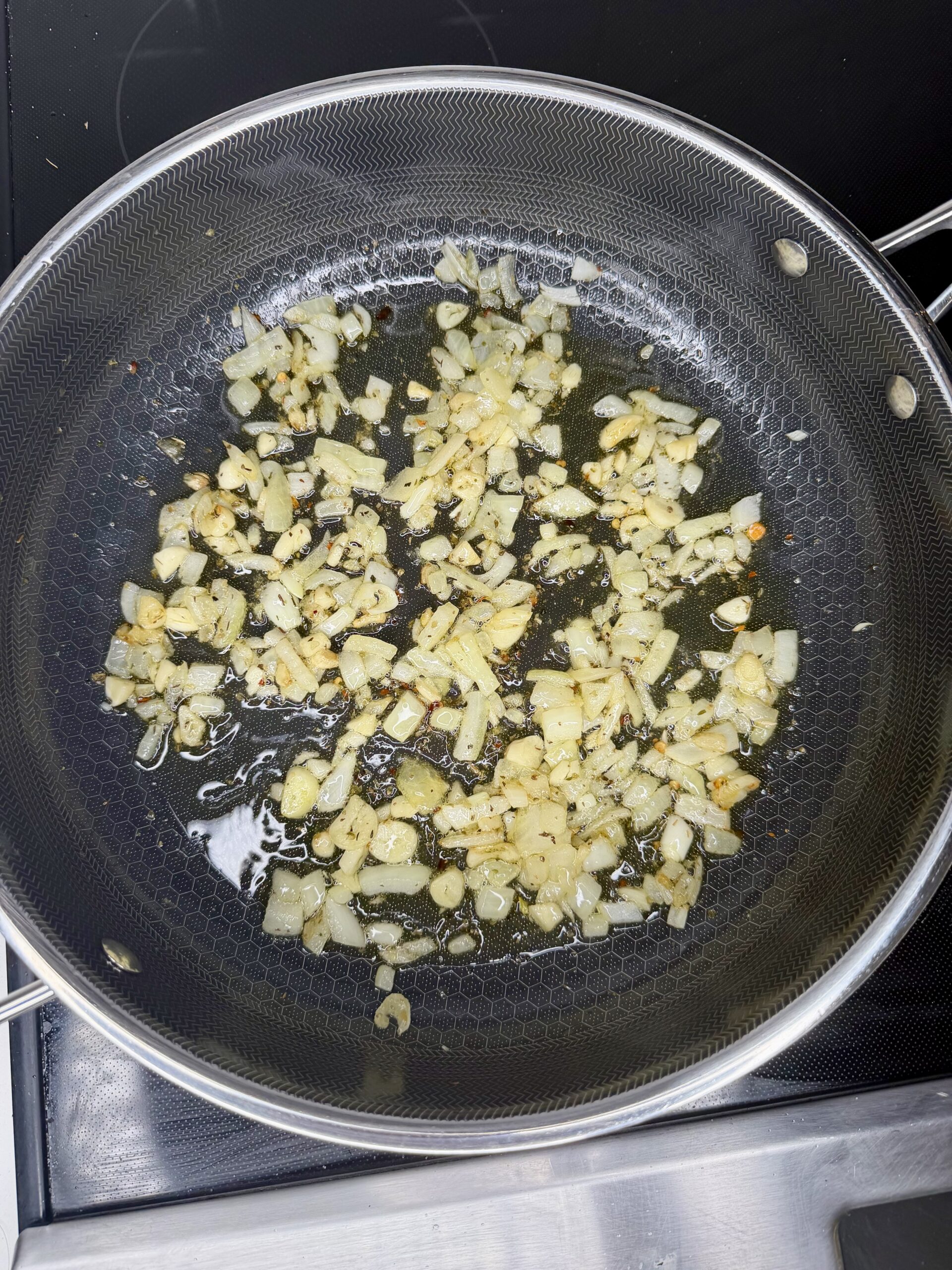 Chopped onions and garlic are sautéing in oil in a frying pan on a stove, beginning to turn golden brown.