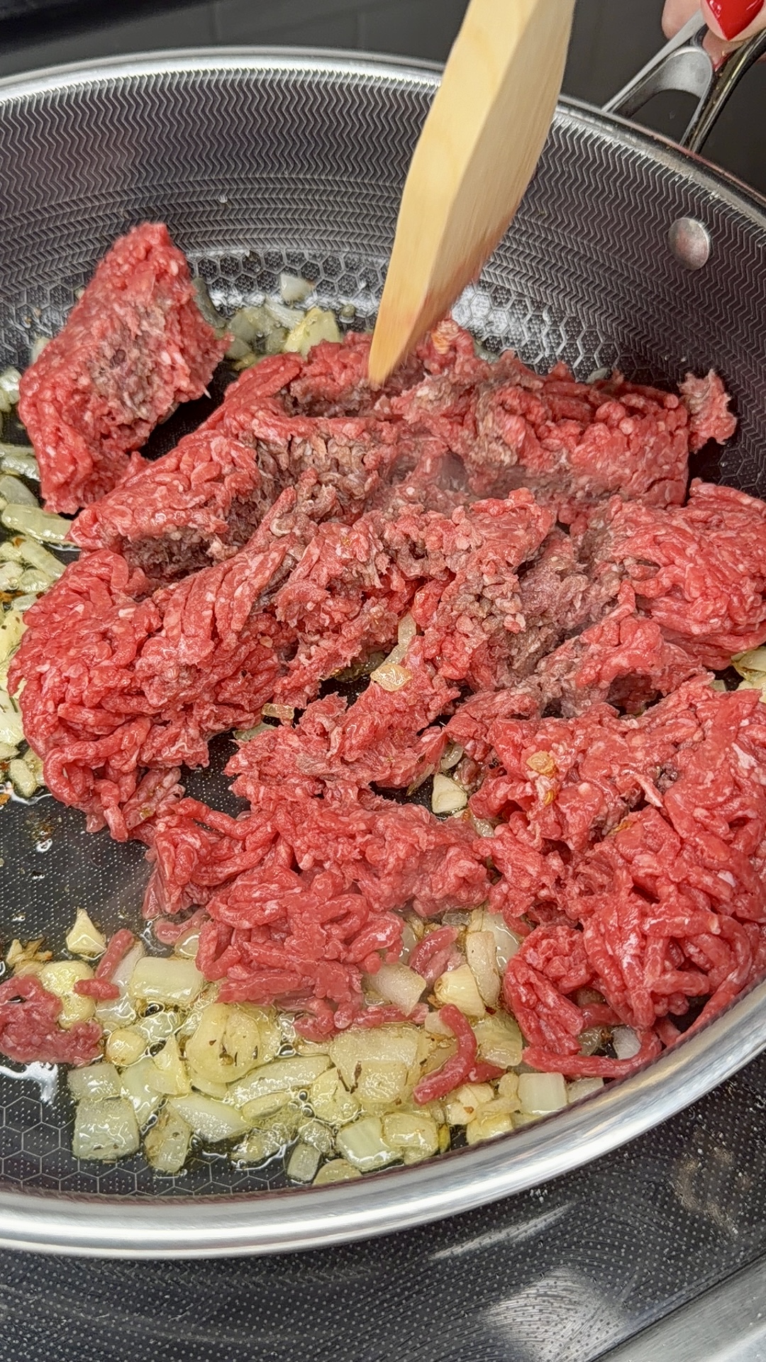 Ground beef and chopped onions cooking in a skillet, with a wooden spatula stirring the meat, starting to brown and mix the ingredients.