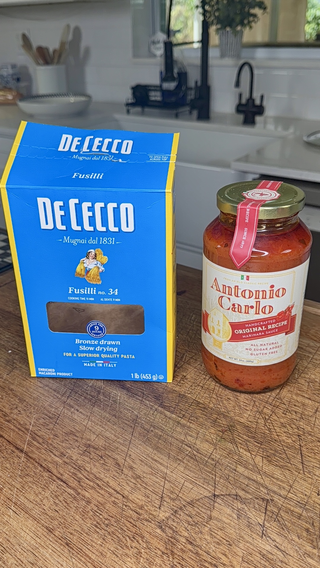 A box of De Cecco Fusilli pasta and a jar of Antonio Carlo tomato basil pasta sauce sit side by side on a wooden kitchen counter with a modern kitchen in the background.