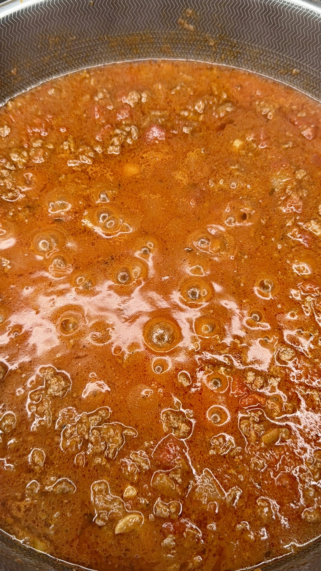 A close-up of a simmering pot of ground beef and tomato sauce, showing a rich, red, oily surface with visible meat and bubbles forming on top.