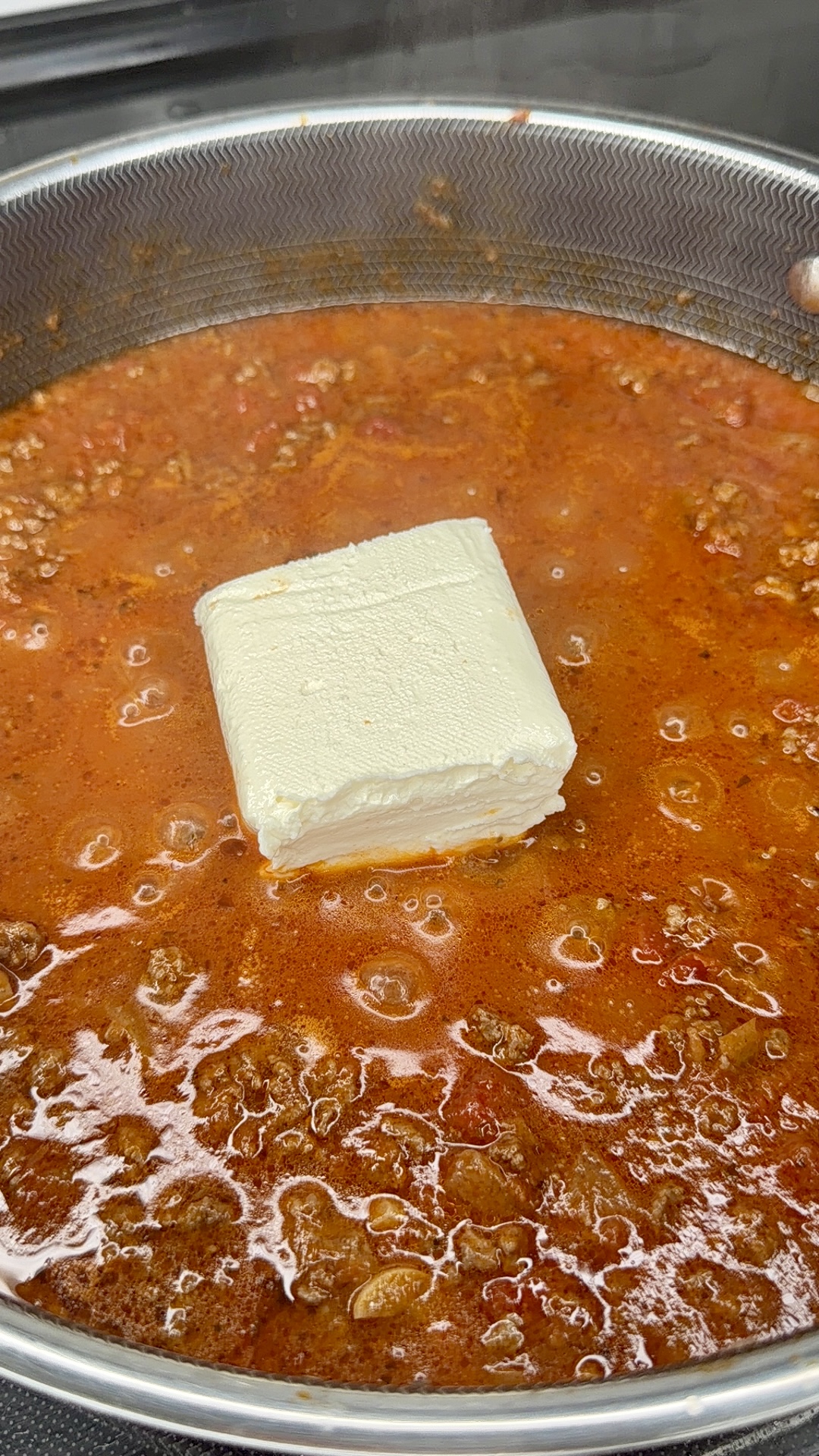 Ground beef simmering in a pan with tomato sauce, topped with a block of cream cheese that is beginning to melt into the mixture.