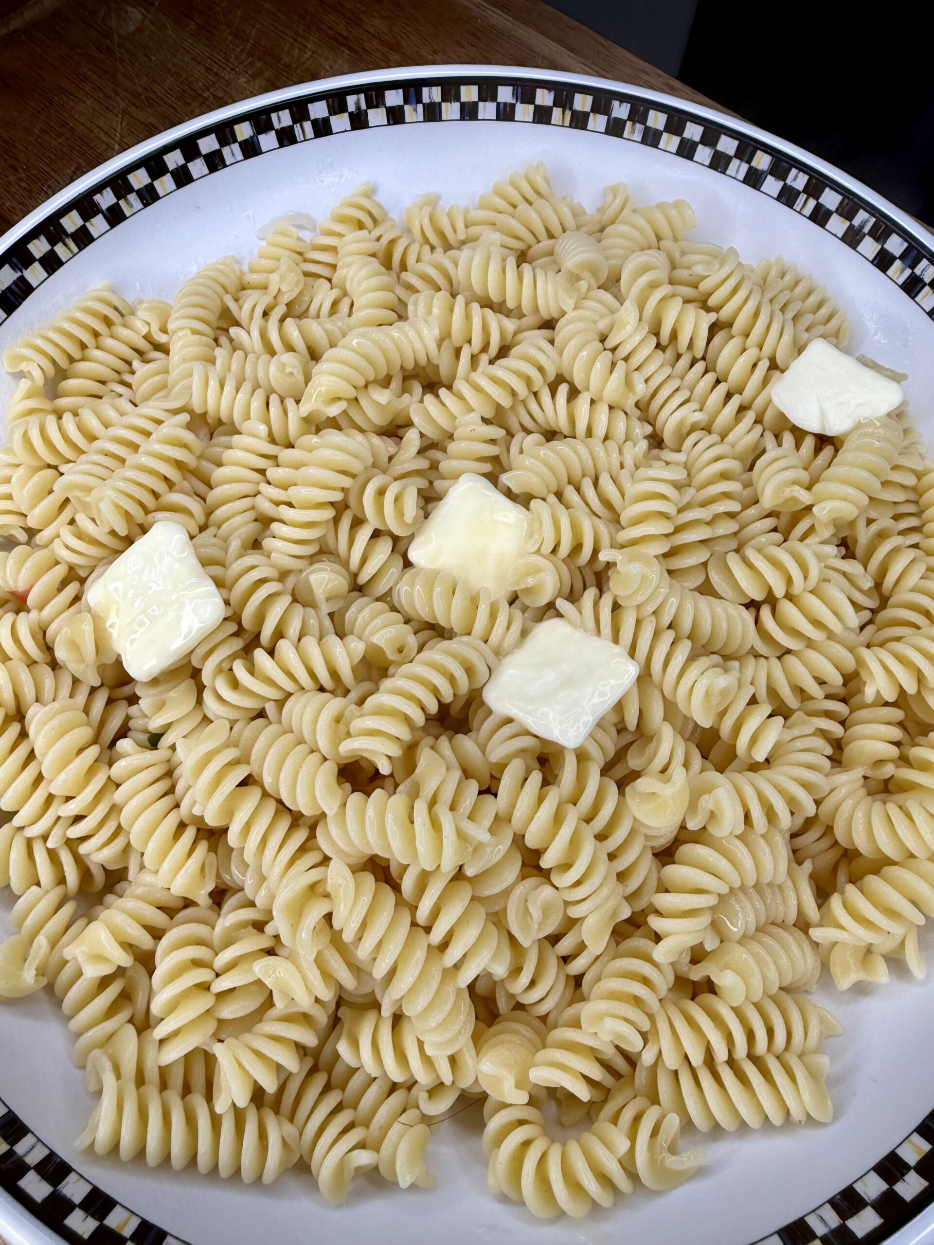 A plate of plain rotini pasta topped with three small squares of butter, on a white dish with a black and white checkered rim.