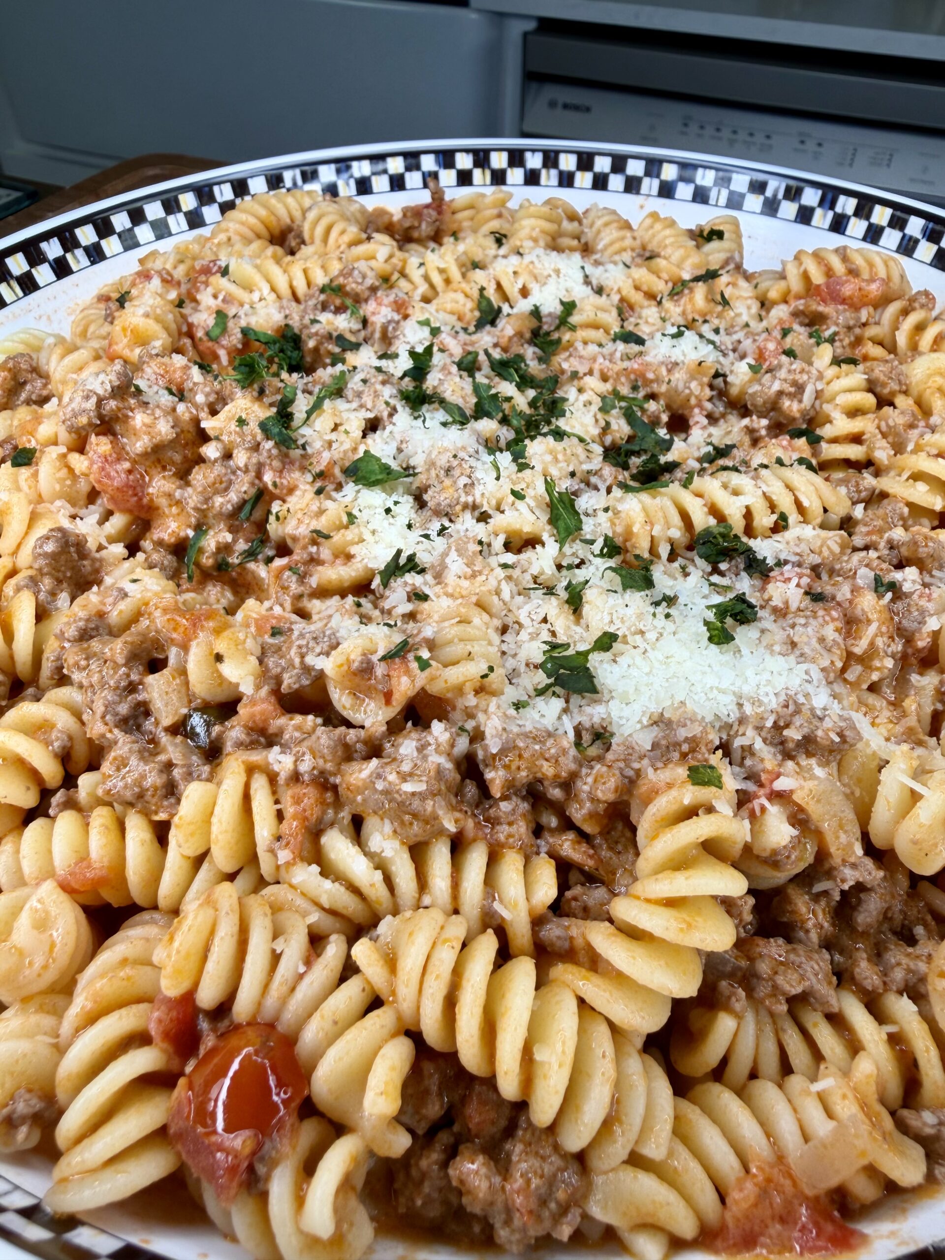 A close-up of a plate of rotini pasta mixed with ground meat, tomato sauce, and topped with grated cheese and chopped parsley. The dish is served in a patterned bowl on a kitchen counter.