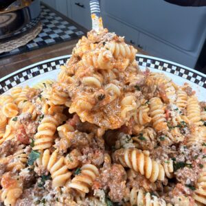 A close-up of a hand holding a serving spoonful of rotini pasta with ground meat, tomato sauce, and herbs over a large plate filled with the same pasta dish. A pot sits in the background on a checkered countertop.