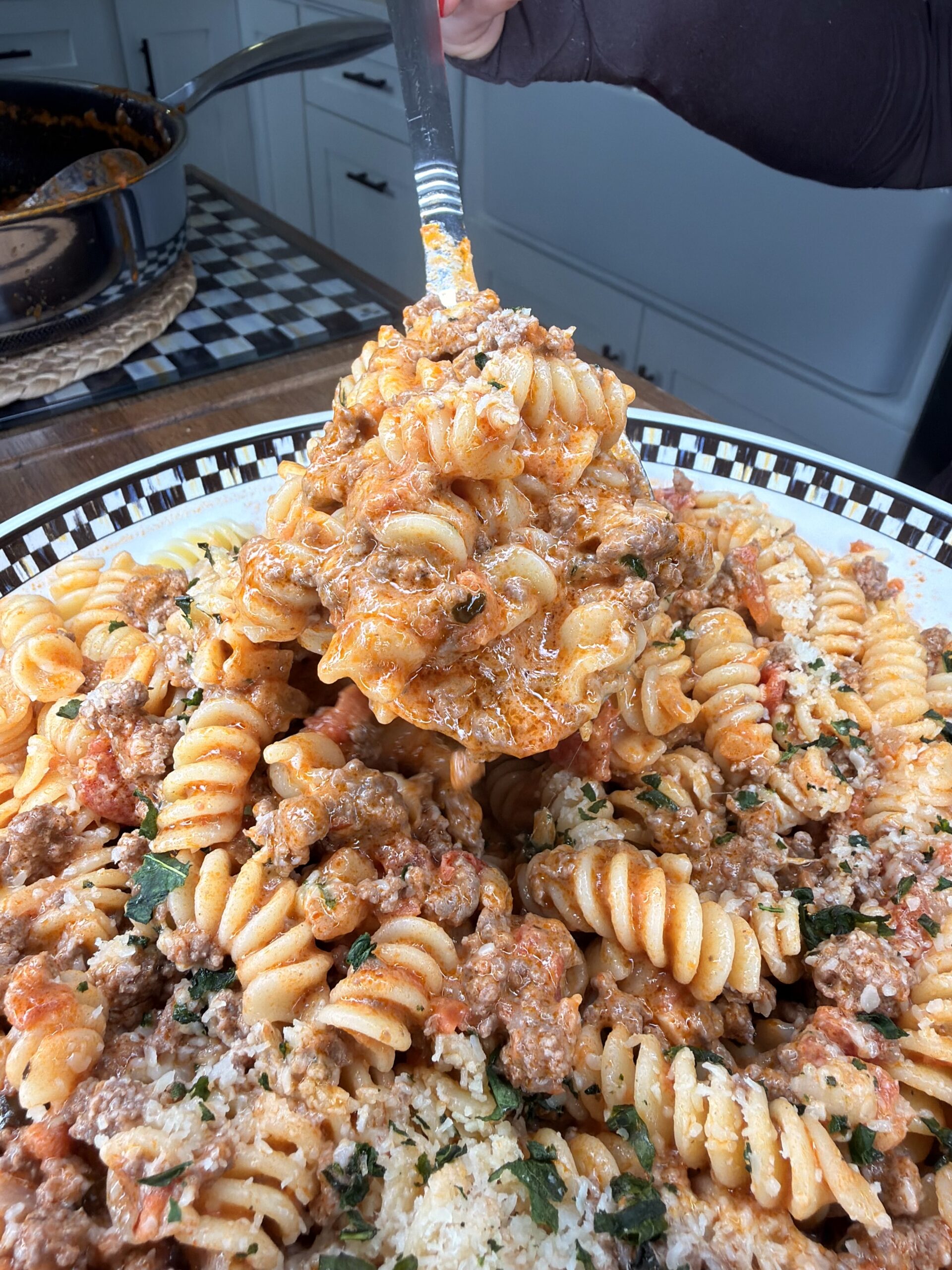 A close-up of a person serving rotini pasta with meat sauce and cheese from a large patterned bowl, with a pot of sauce and kitchen counter visible in the background.