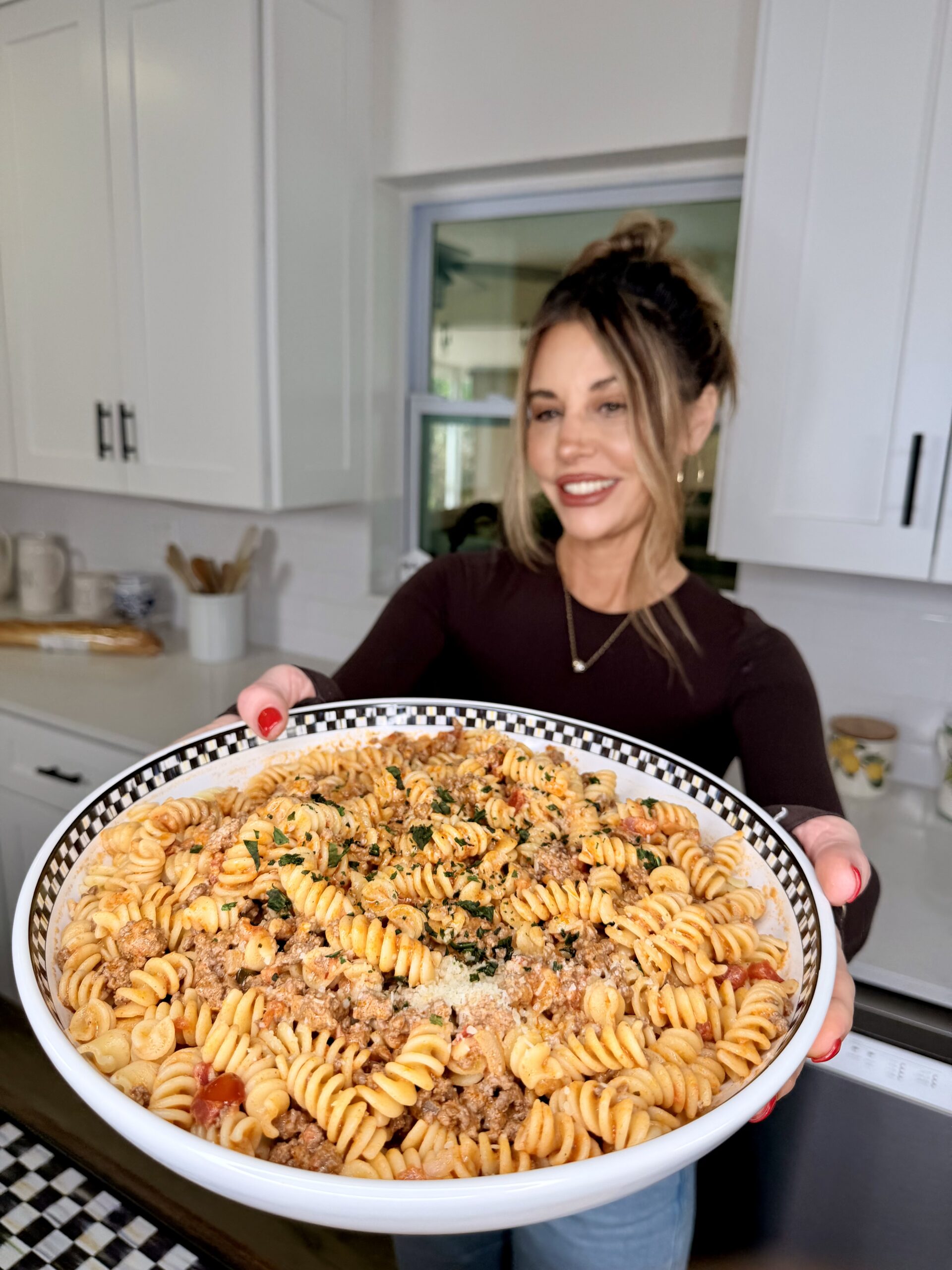 A smiling woman in a kitchen holds out a large bowl of rotini pasta topped with ground meat, tomato sauce, and herbs. White cabinets and a window are visible in the background.