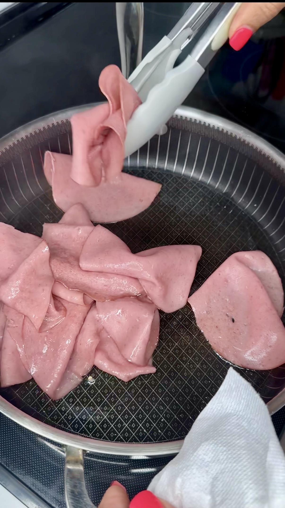 Slices of pink bologna are being placed into a frying pan with tongs. Some slices are already frying and starting to curl at the edges. A hand with red-painted nails is holding the tongs.