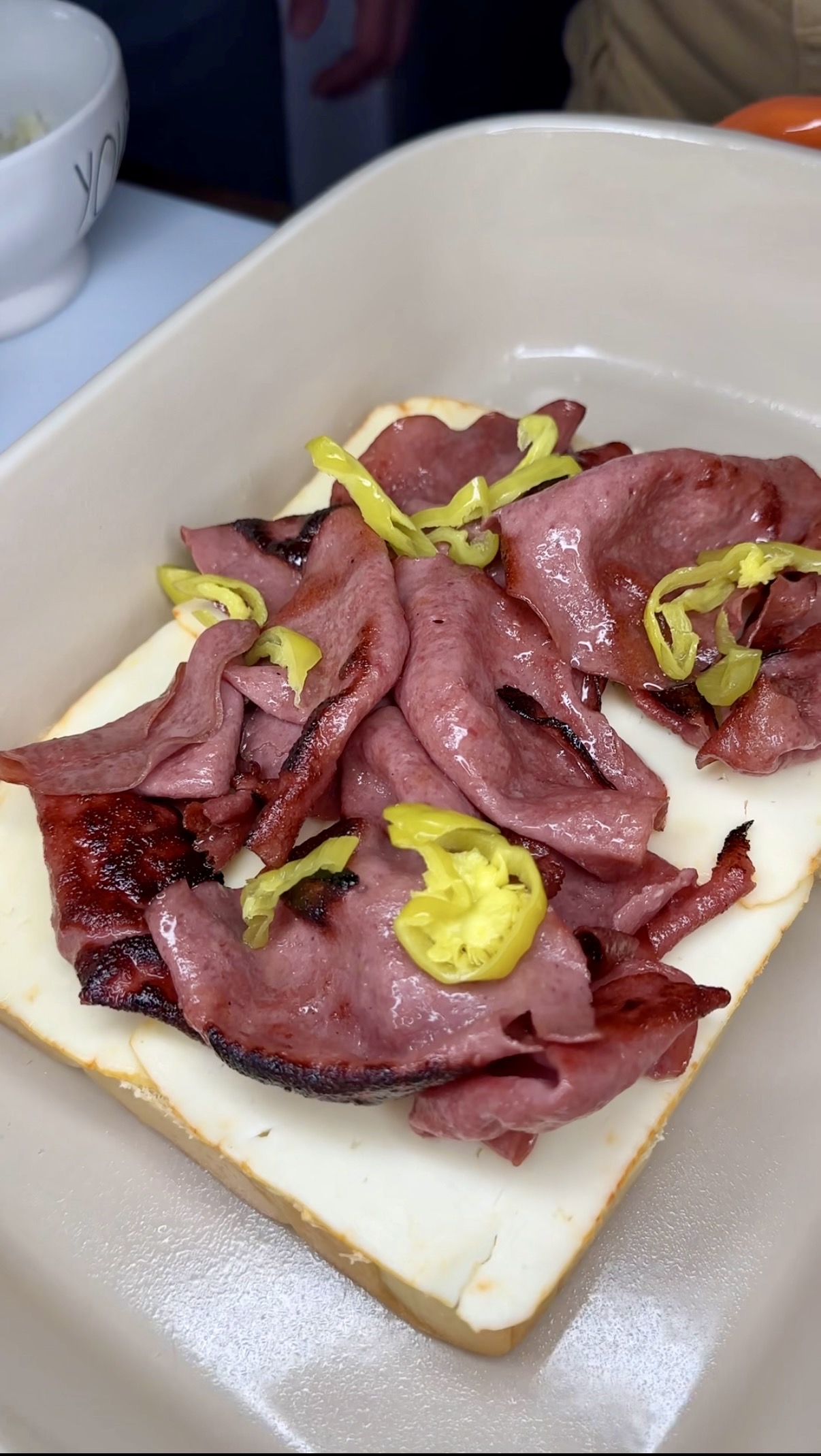 An open-faced sandwich with melted white cheese, slices of grilled pastrami, and yellow pepper rings on top, served on a rectangular piece of bread in a beige dish.