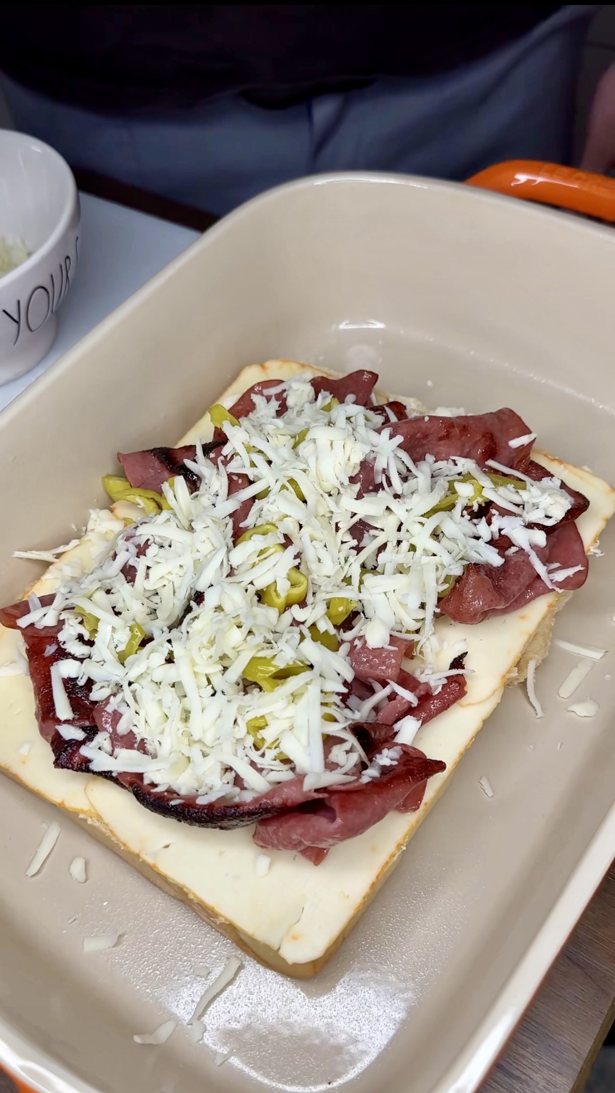 A slice of white bread topped with sliced meat, shredded cheese, and chopped pickles sits in a beige baking dish. A portion of a white bowl is visible in the background.