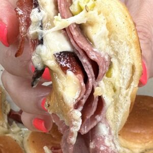 A hand with pink-painted nails holds a sandwich filled with melted cheese, sliced meats, sauerkraut, and peppers, with buns visible in the background.