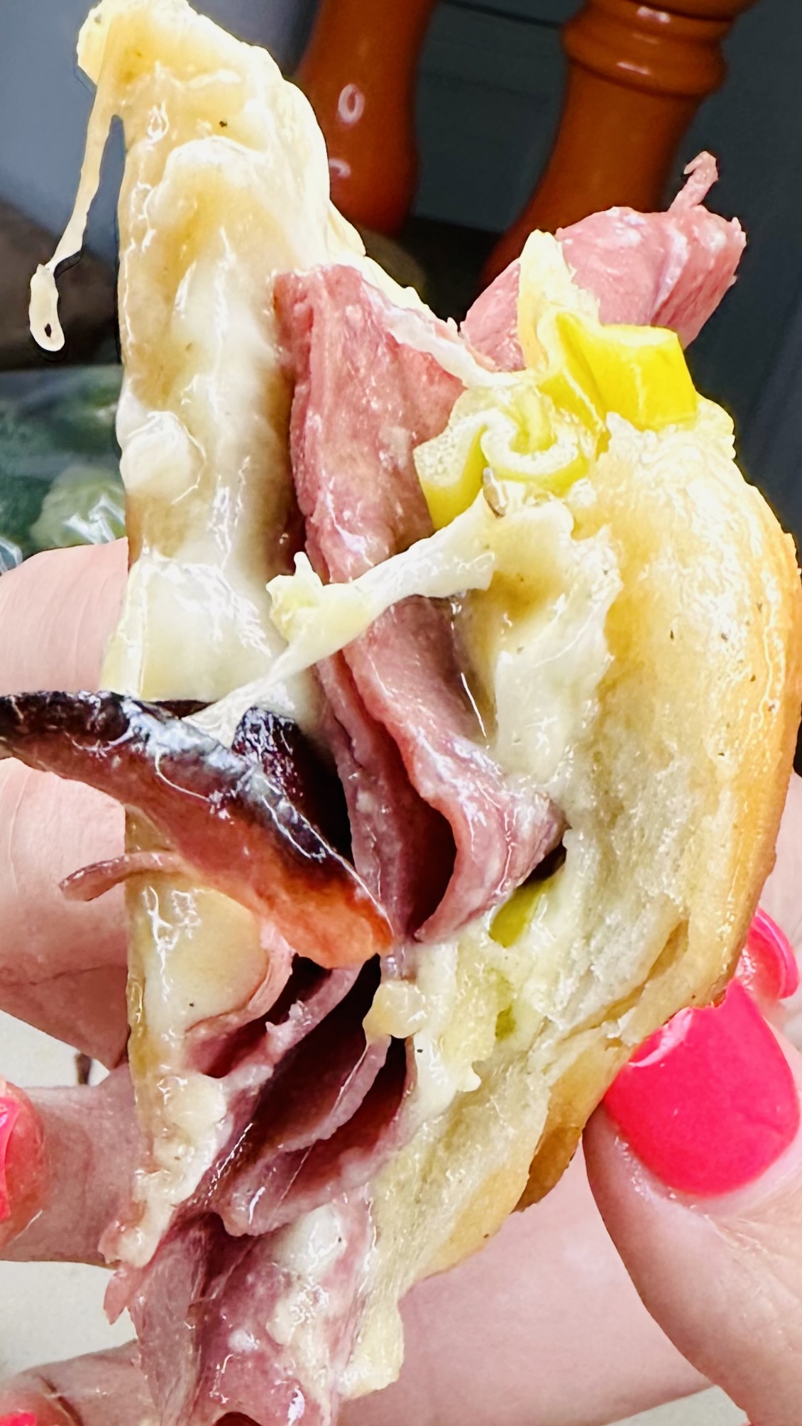 A close-up of a hand holding a sandwich filled with melted cheese, sliced deli meat, pickles, and olives. The person has bright pink painted fingernails.