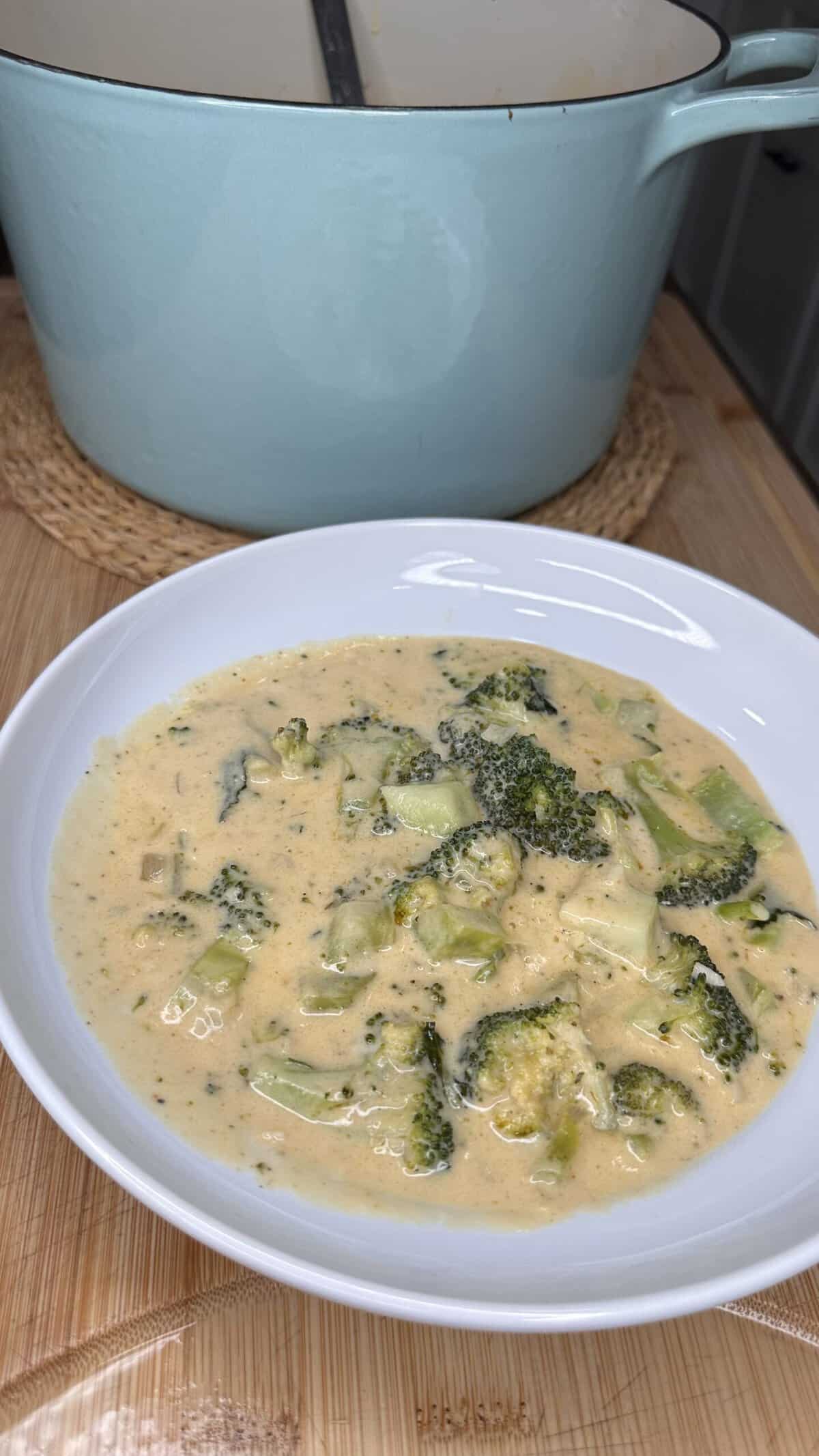 A white bowl filled with creamy broccoli cheddar soup sits on a wooden surface. In the background, there is a large light blue pot on a woven trivet.