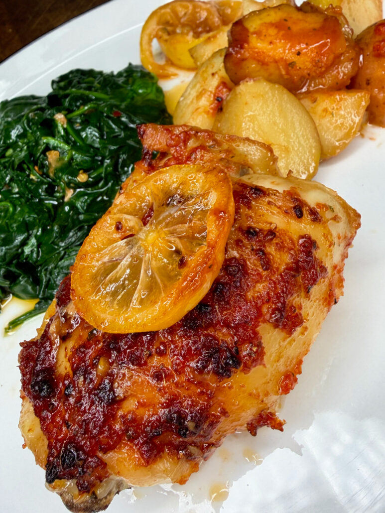 A plate of Italian Roast Chicken and Potatoes, baked to perfection and topped with a lemon slice, is served alongside sautéed spinach, with roasted carrots adding color in the background.