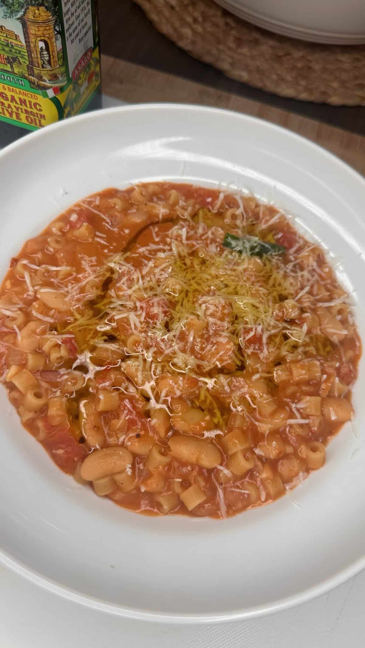 A white bowl filled with pasta e fagioli soup, featuring ditalini pasta, white beans, tomato broth, greens, a drizzle of olive oil, and grated cheese on top.