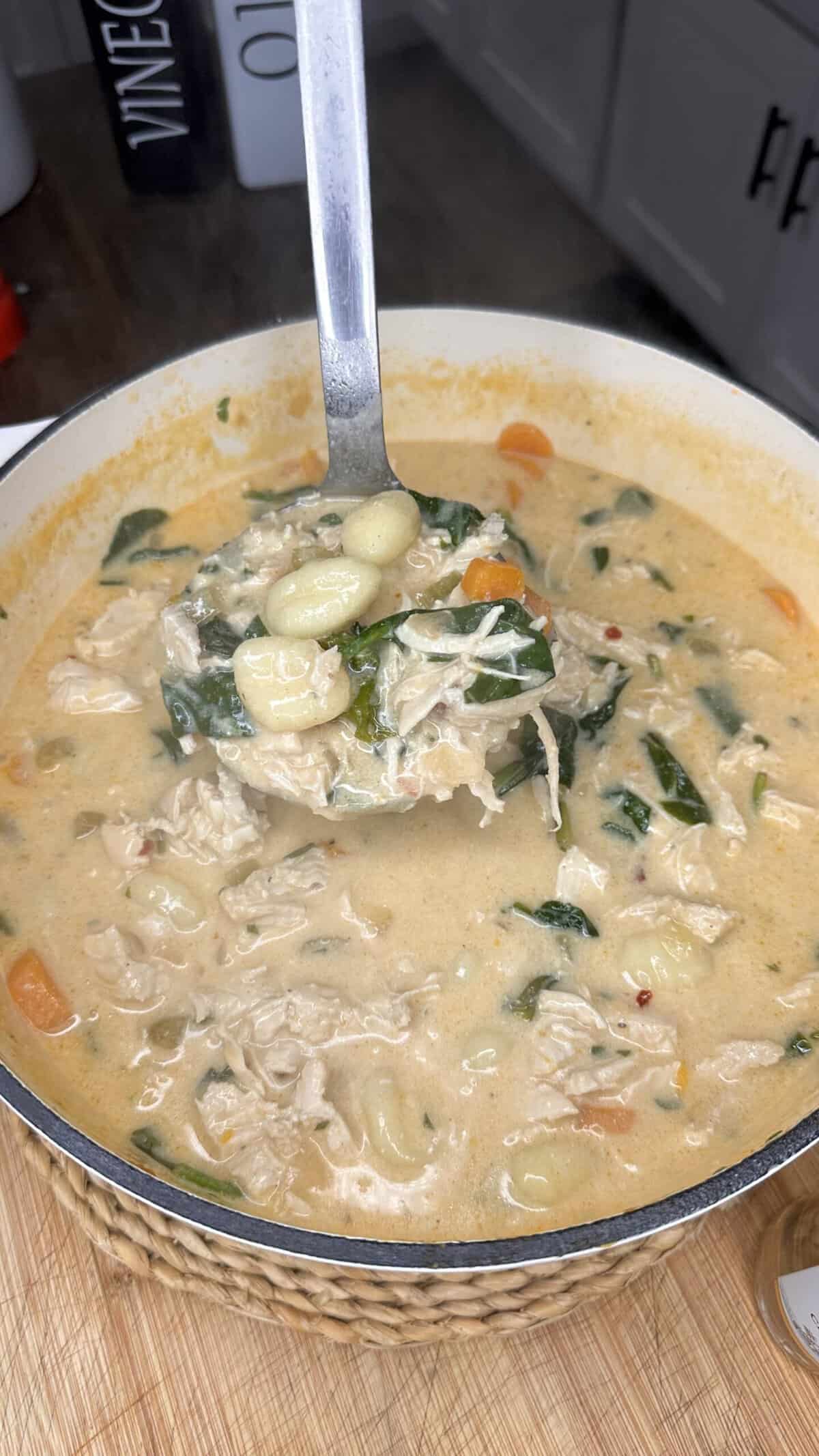 A ladle lifts creamy chicken gnocchi soup with spinach, carrots, and shredded chicken from a large pot on a kitchen counter.