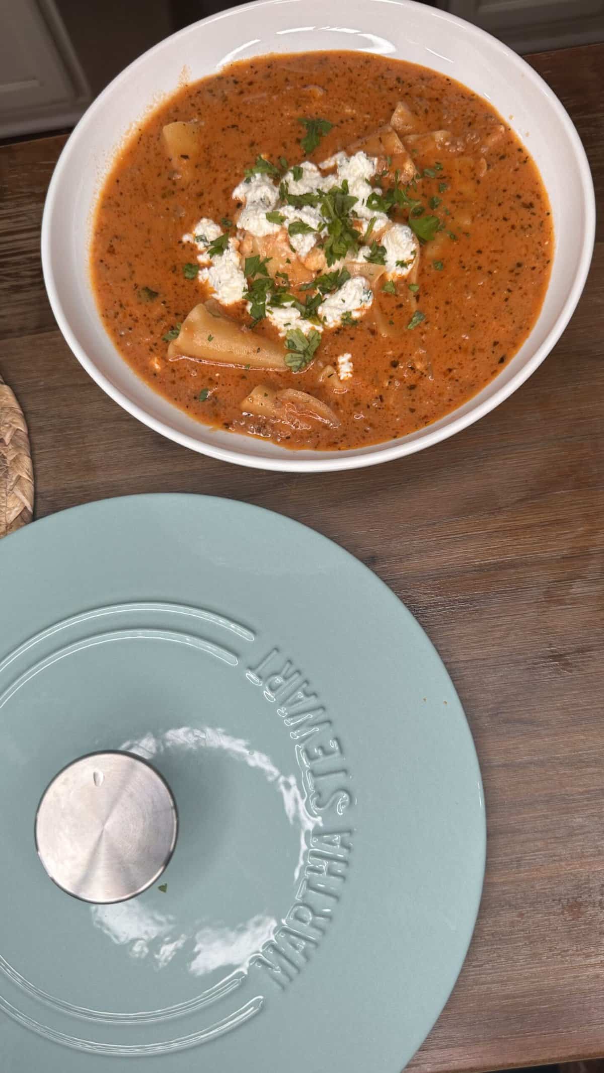 A bowl of tomato-based soup with pasta, crumbled cheese, and chopped herbs sits on a wooden surface next to the lid of a Martha Stewart cookware pot.