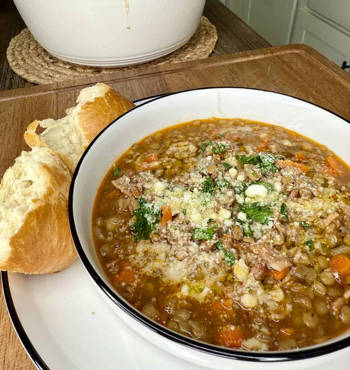 A bowl of lentil soup with ground meat, carrots, and herbs, topped with grated cheese, served with two slices of crusty bread on a plate. A white pot sits in the background on a wooden surface.