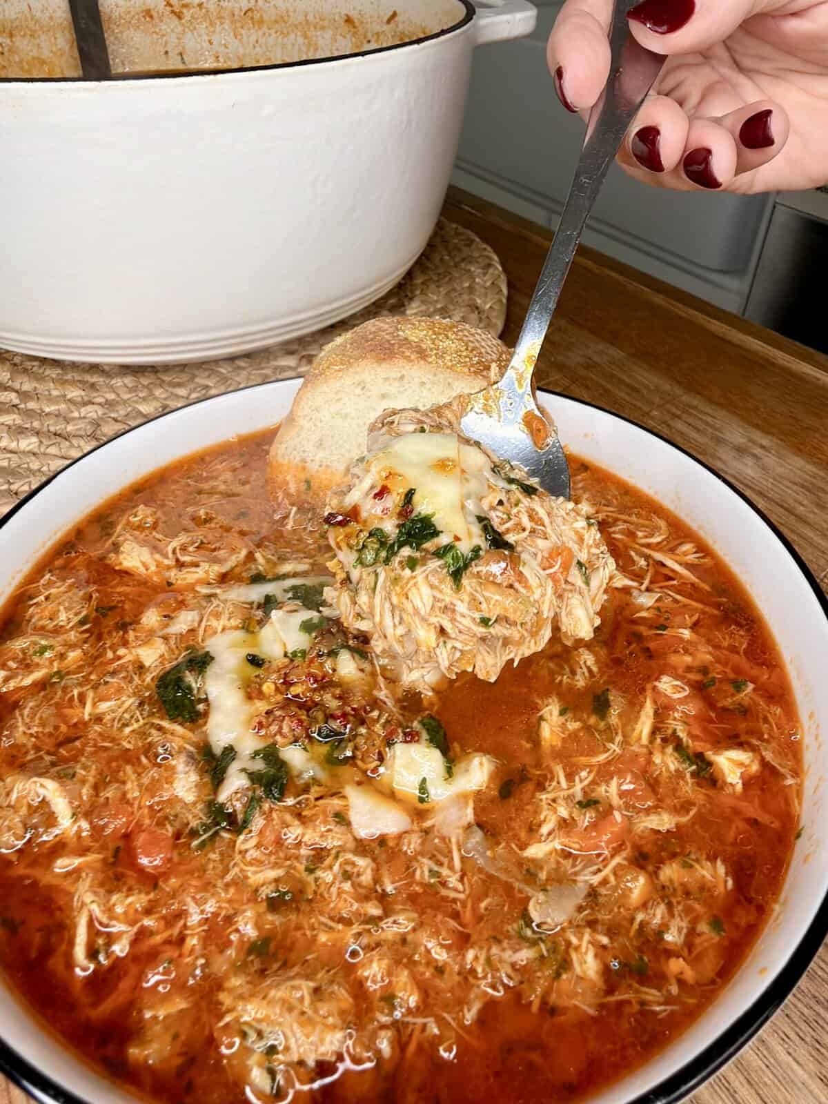 A bowl of shredded chicken soup in a rich red broth topped with melted cheese and herbs. A hand holds a spoonful of the soup above the bowl, with a slice of bread resting on the edge. A white pot is in the background.