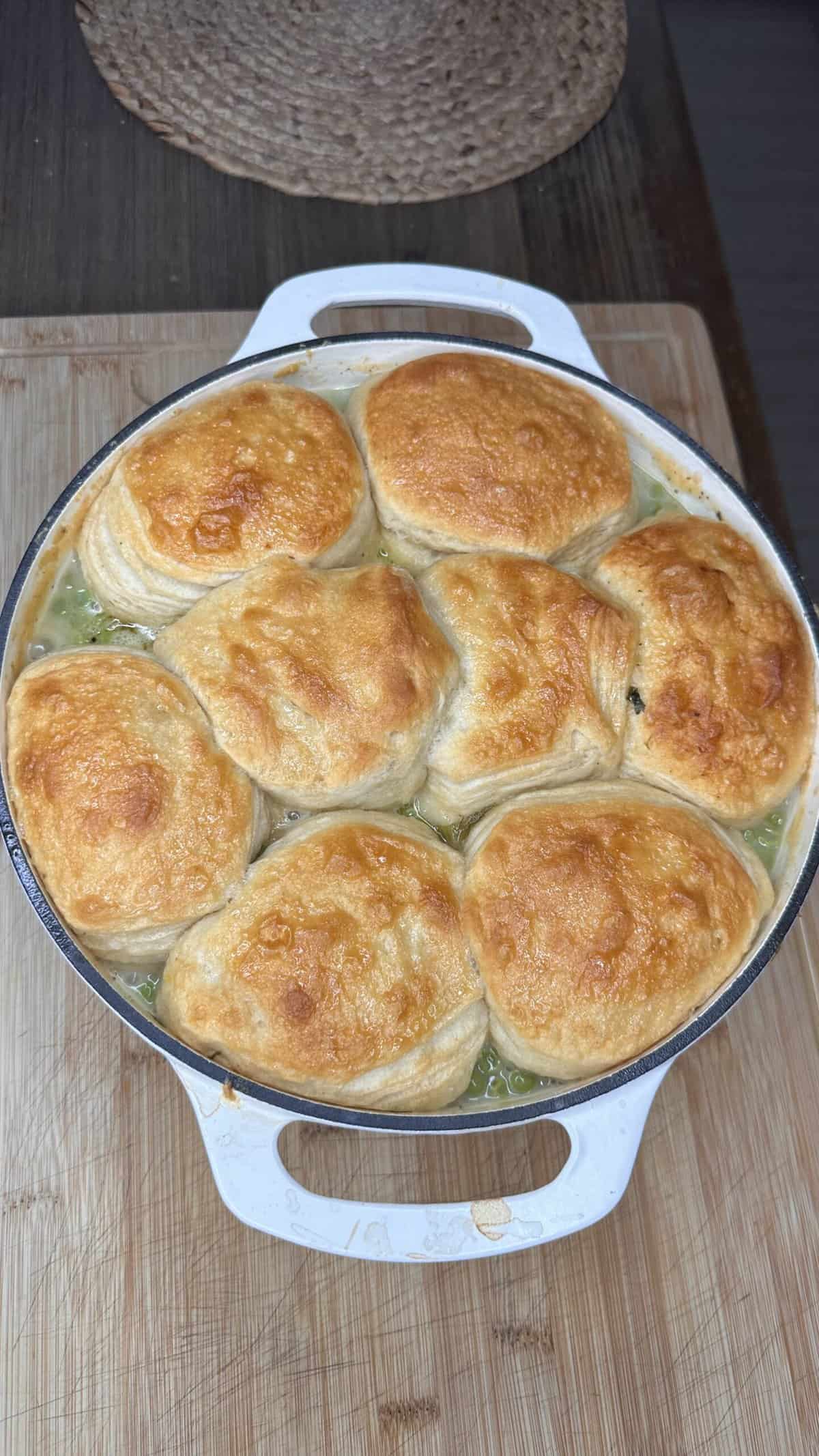 A round white casserole dish filled with creamy pot pie topped with eight golden-brown, flaky biscuits, sitting on a wooden cutting board.