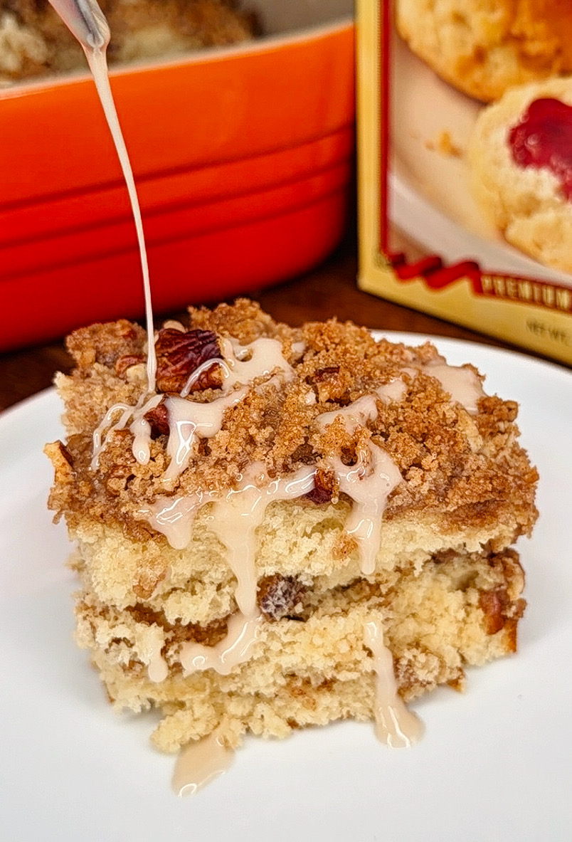 A slice of crumb cake with raisins and pecans sits on a white plate while icing is being drizzled on top. A red baking dish and a box with a biscuit image are visible in the background.