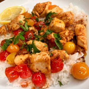 A plate of white rice topped with cooked chicken pieces, red and yellow cherry tomatoes, chopped parsley, and a lemon wedge on the side.