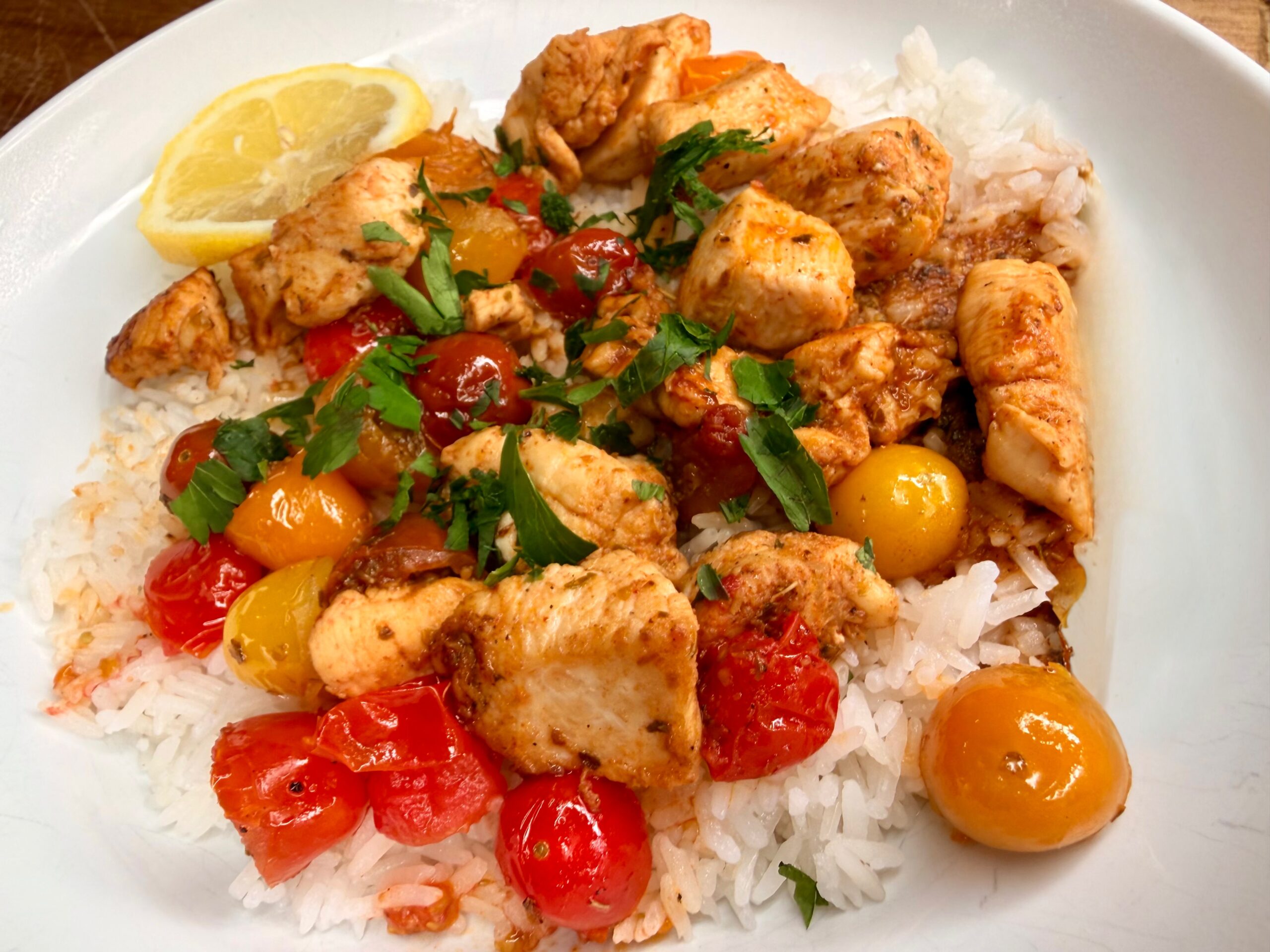 A plate of white rice topped with cooked chicken pieces, red and yellow cherry tomatoes, chopped parsley, and a lemon wedge on the side.