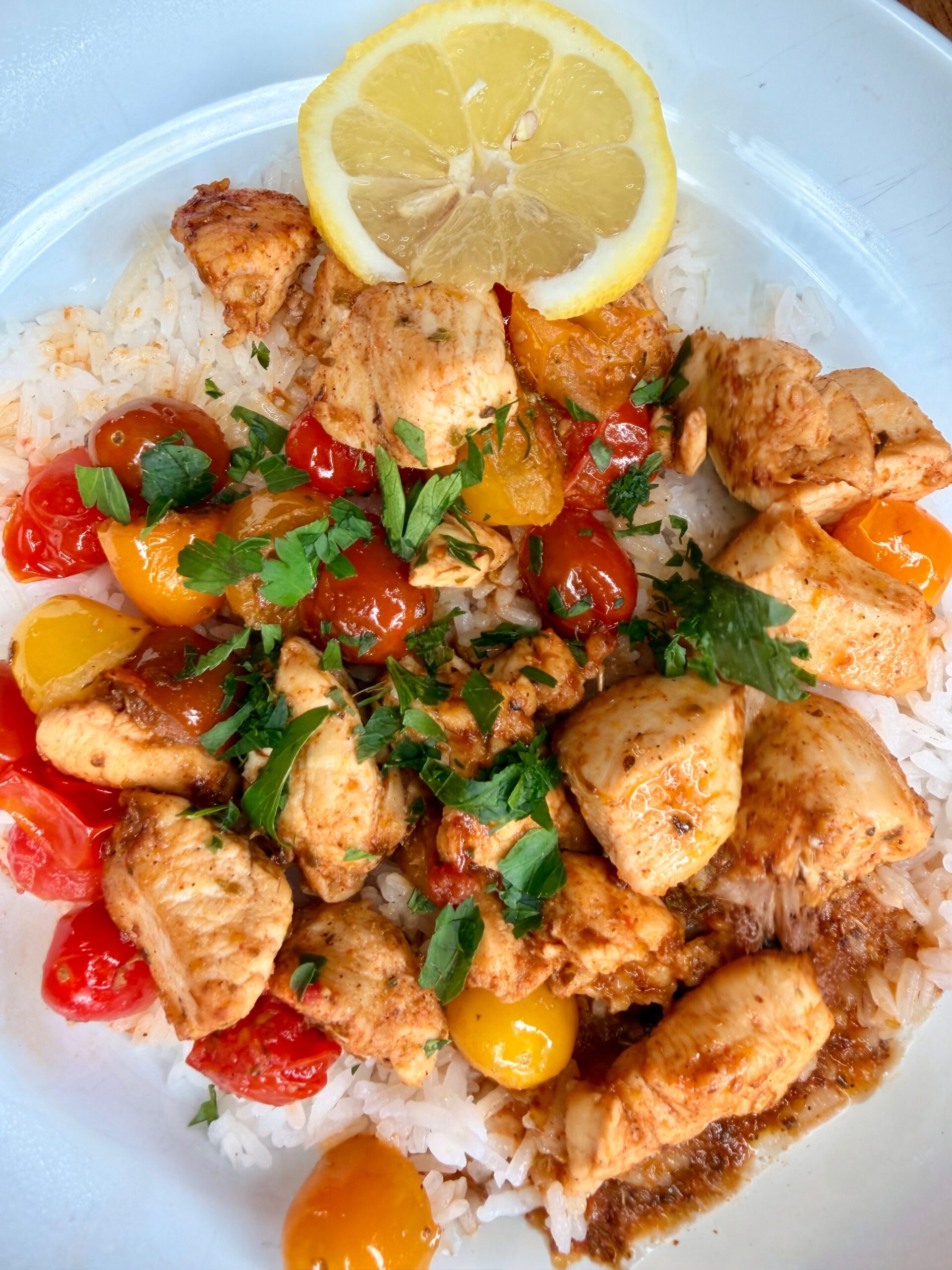 A plate of white rice topped with seasoned chicken pieces, red and yellow cherry tomatoes, chopped herbs, and garnished with a lemon slice.