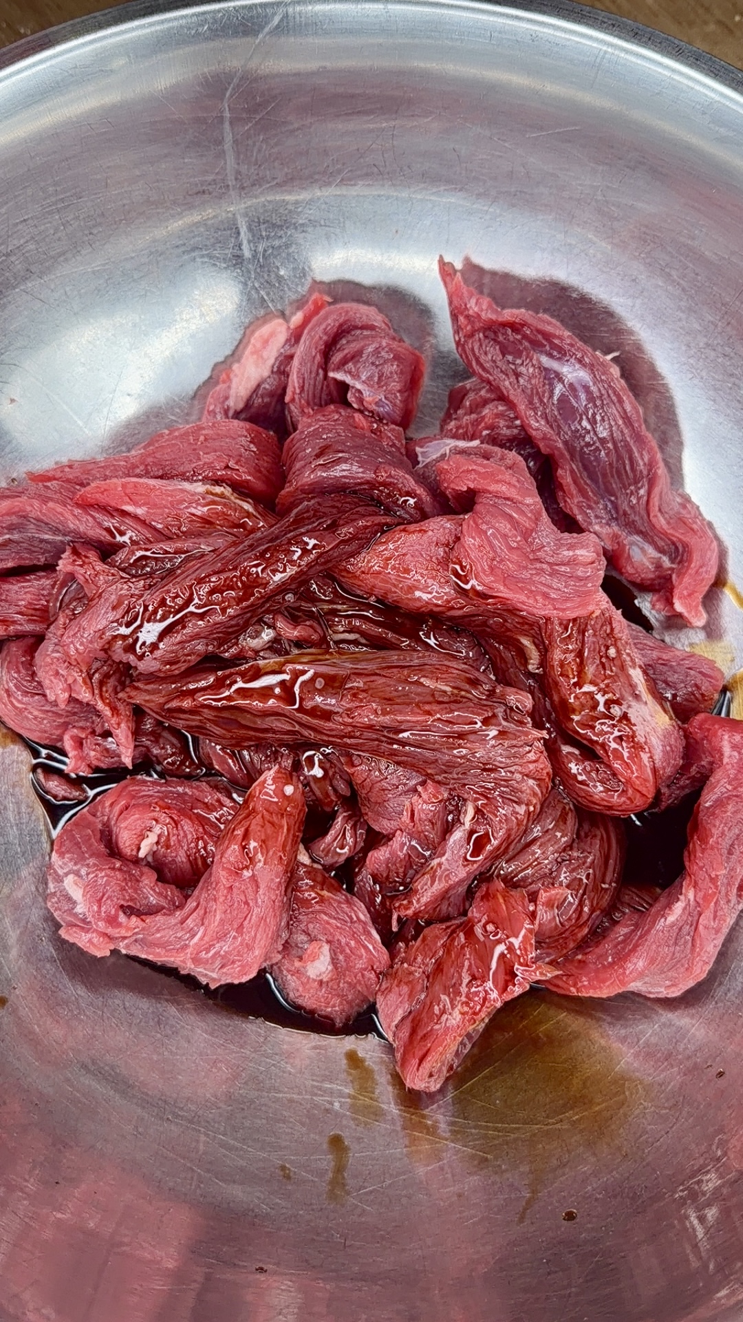 Raw slices of red beef marinating in a metal bowl, coated with a glossy liquid.