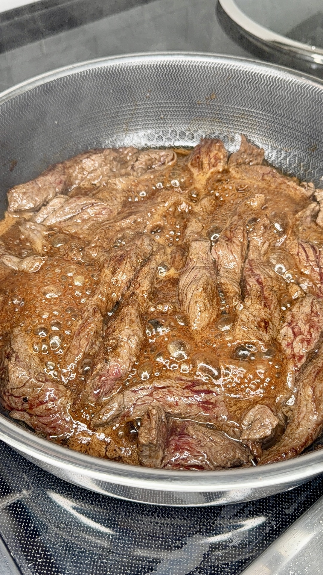 Strips of beef are sizzling and browning in a pan filled with bubbling sauce on a stovetop.