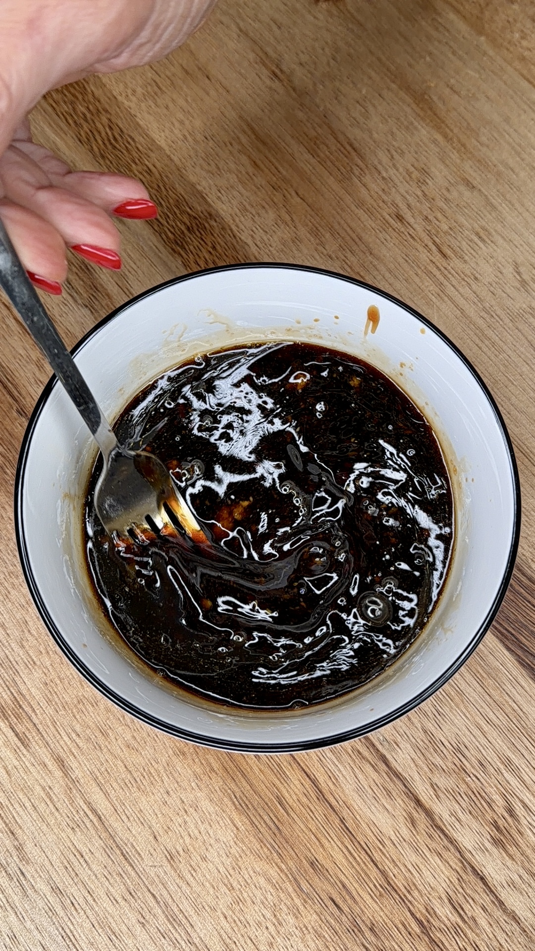 A hand with red nail polish holds a spoon, stirring a dark, glossy sauce in a white bowl with a black rim on a wooden surface.