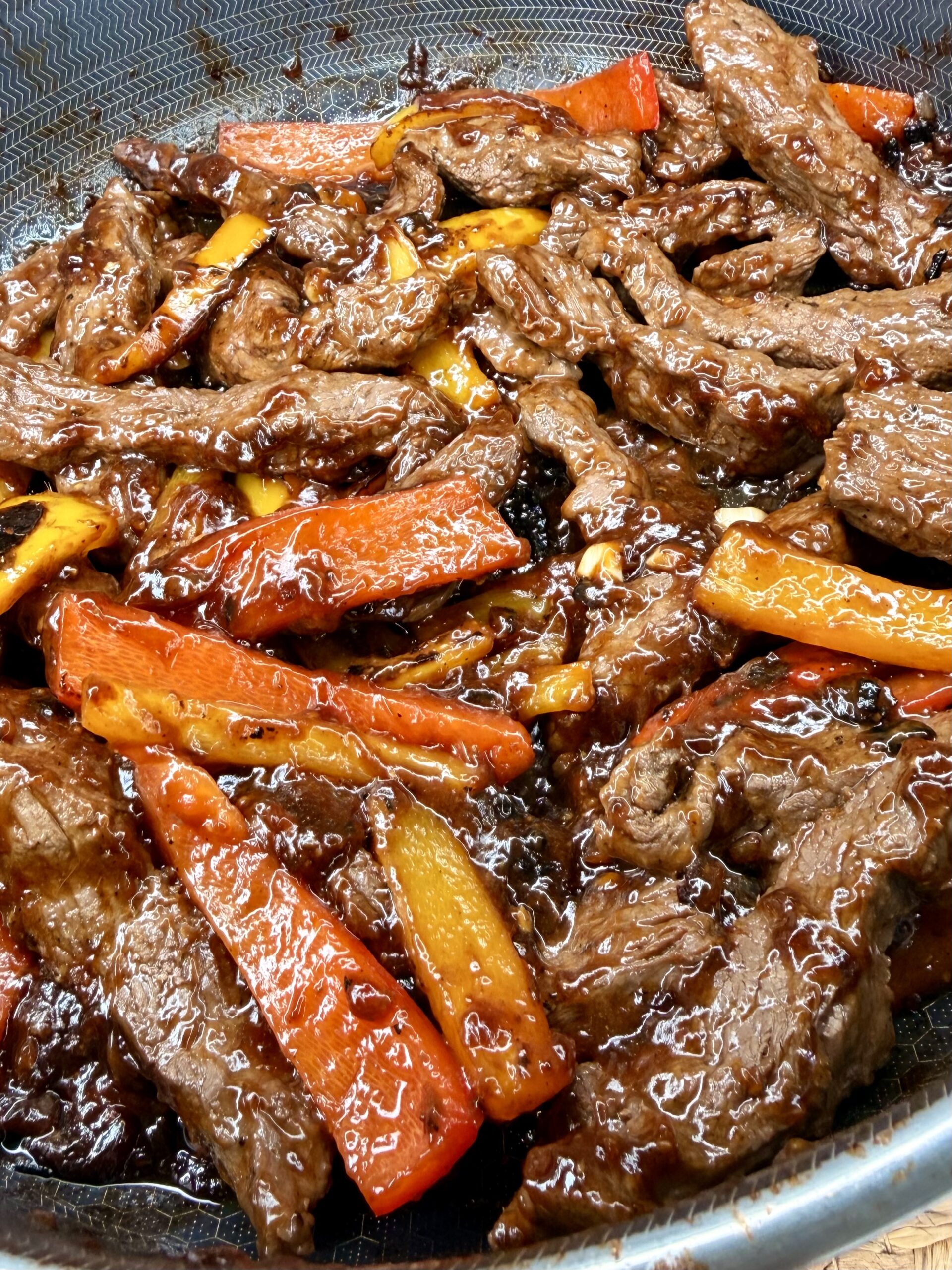 Sliced beef strips and red and yellow bell pepper strips cooked in a glossy brown sauce, all in a skillet, creating a savory stir-fry dish.