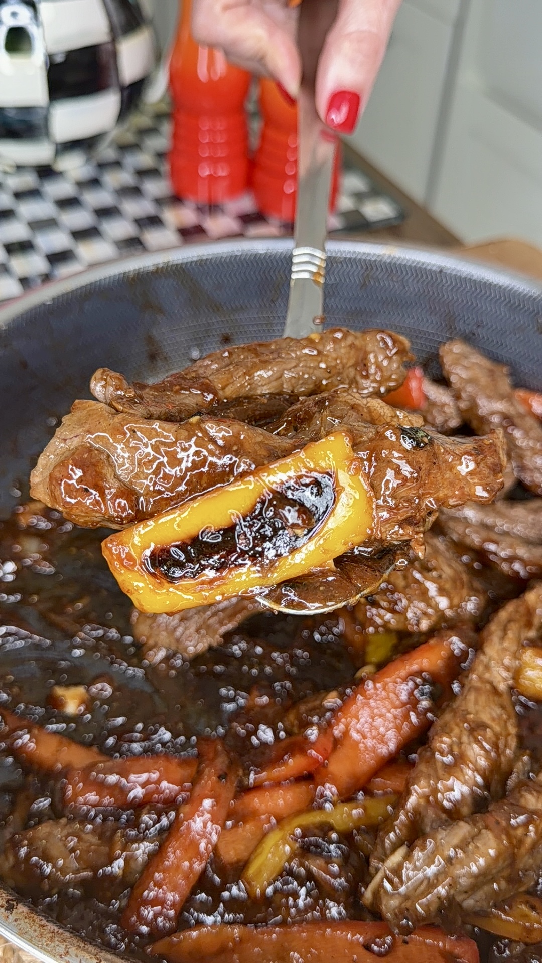 A close-up of a spoon lifting sautéed beef strips, yellow bell pepper, and carrots coated in a glossy brown sauce from a skillet. A hand with red nail polish holds the spoon over the pan.