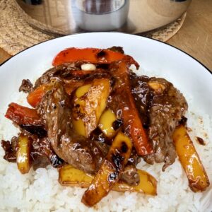 A plate of white rice topped with stir-fried beef strips, red and yellow bell peppers, and a glossy brown sauce. A pan with more of the same stir-fry is visible in the background.