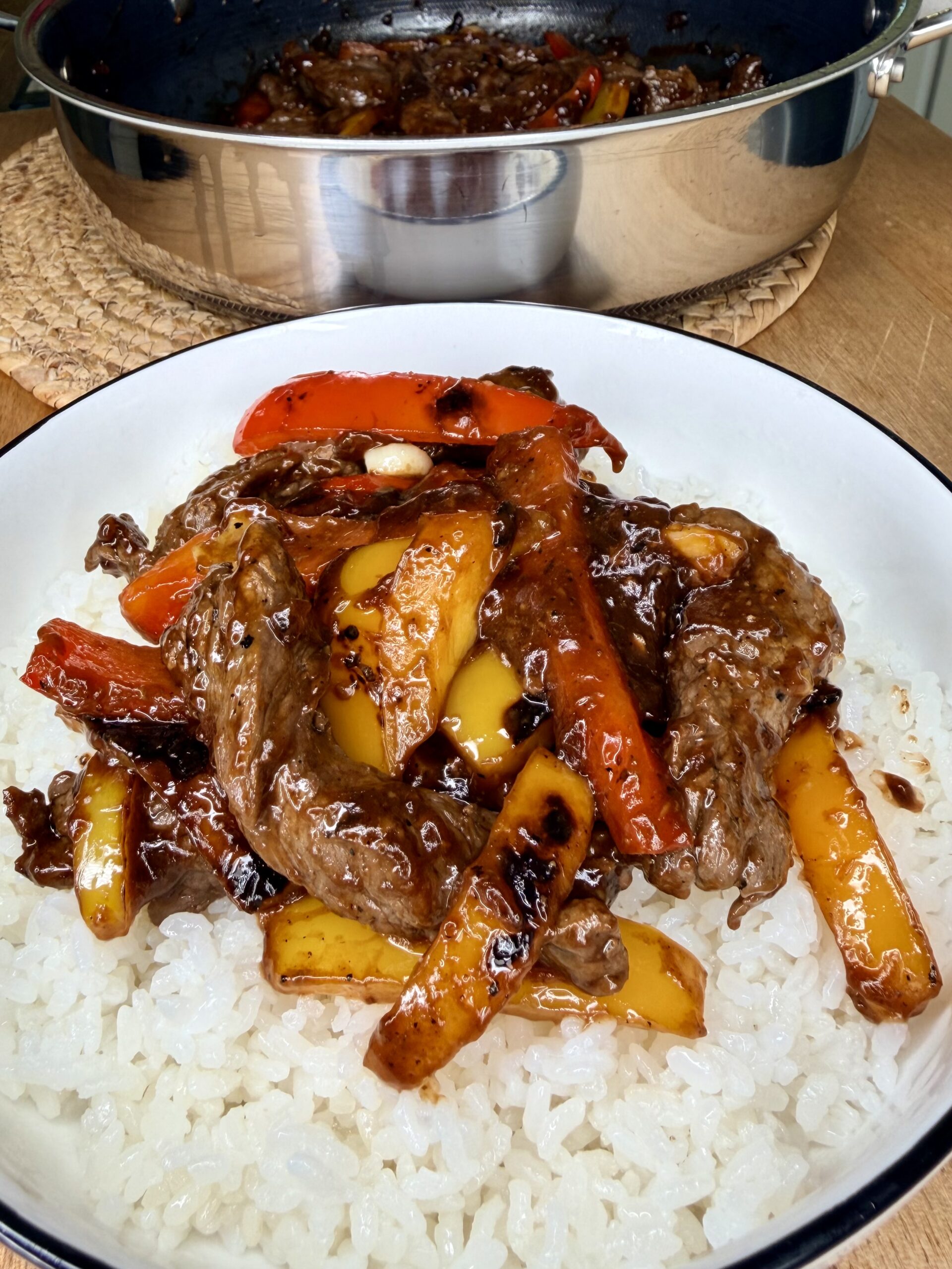 A plate of white rice topped with stir-fried beef strips, red and yellow bell peppers, and a glossy brown sauce. A pan with more of the same stir-fry is visible in the background.