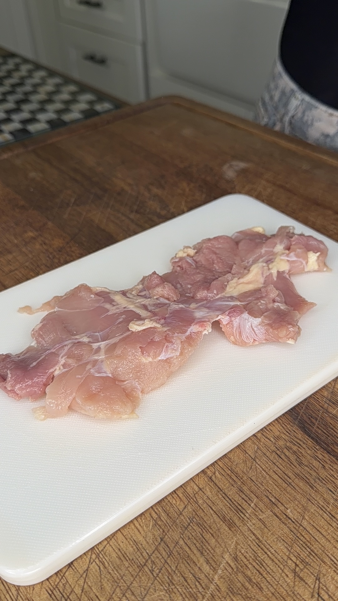 Raw chicken pieces are placed on a white cutting board, which sits on a wooden countertop. The background shows part of a kitchen with cabinets and a patterned floor.