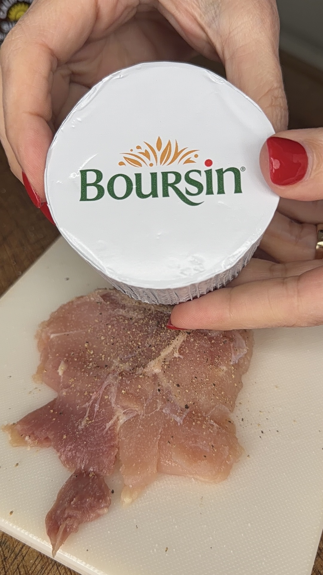 A person with red nail polish holds a container of Boursin cheese above raw, seasoned meat on a white cutting board.