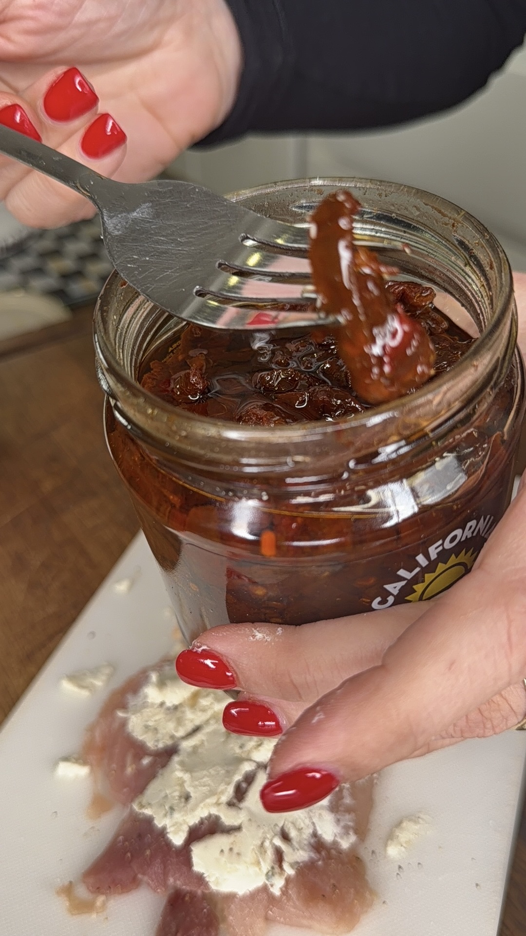 A person with red-painted nails uses a fork to remove sun-dried tomatoes from a jar, with raw chicken breast and cheese visible on a cutting board below.