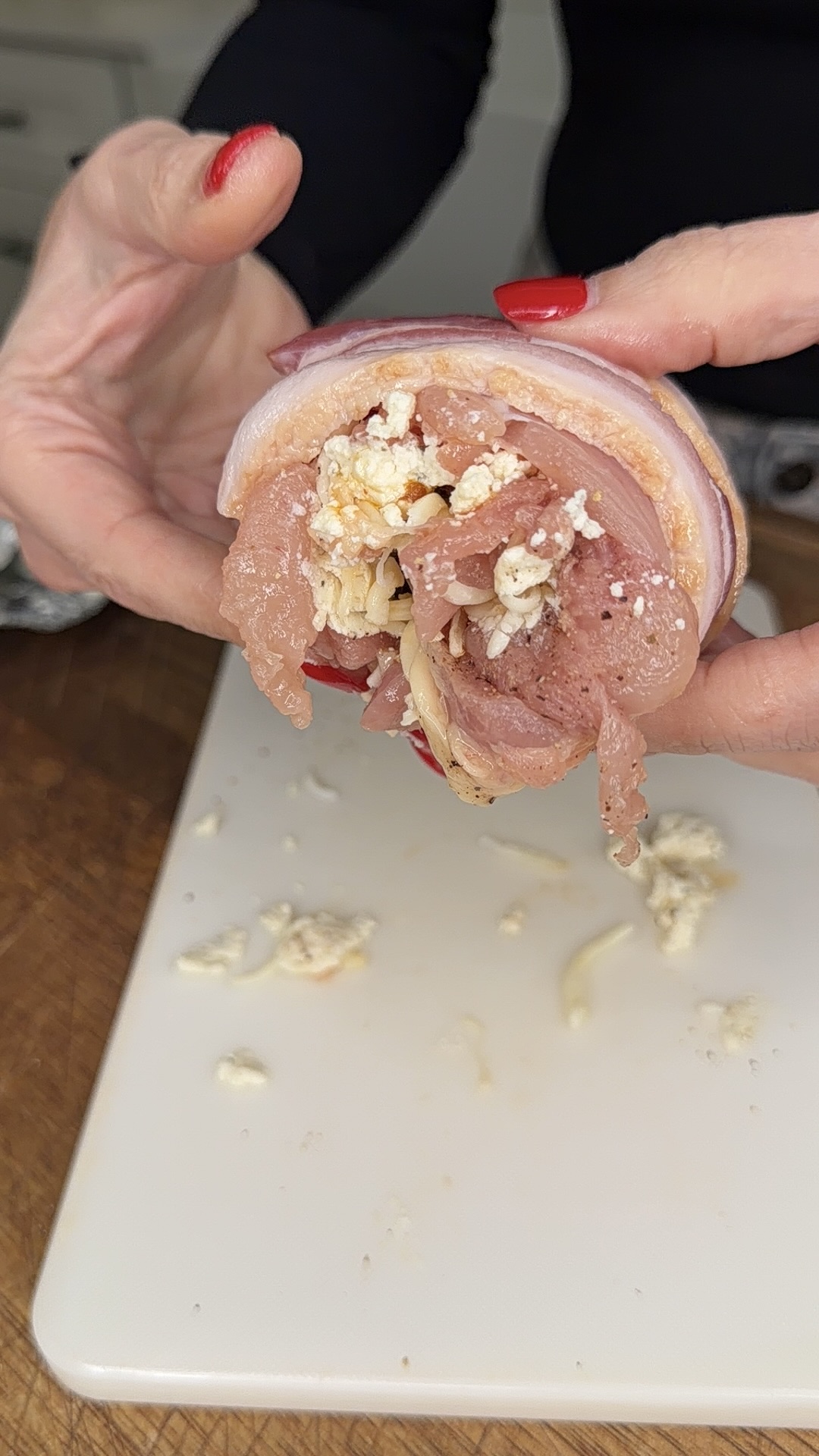 A person with red nail polish holds a partially assembled raw chicken breast stuffed with cheese and spices, wrapped in bacon. A white cutting board with some cheese crumbles is visible below.