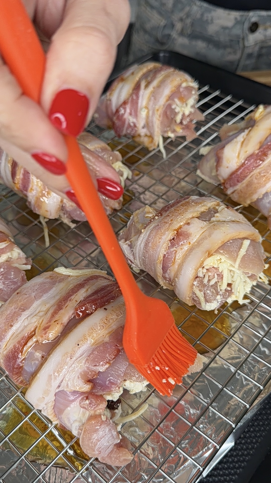 A hand with red nails uses an orange brush to apply sauce to bacon-wrapped chicken pieces, placed on a wire rack over a baking tray.