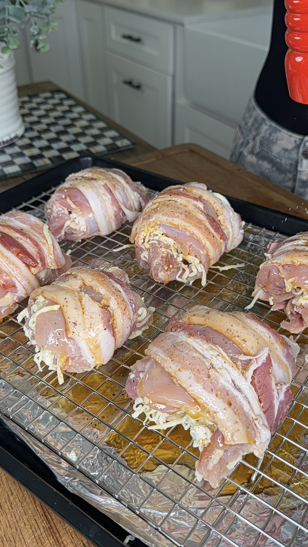 Raw chicken breasts stuffed with shredded cheese and wrapped in bacon are arranged on a wire rack over a baking sheet. A person is sprinkling seasoning or pepper over the prepared chicken. Kitchen background visible.