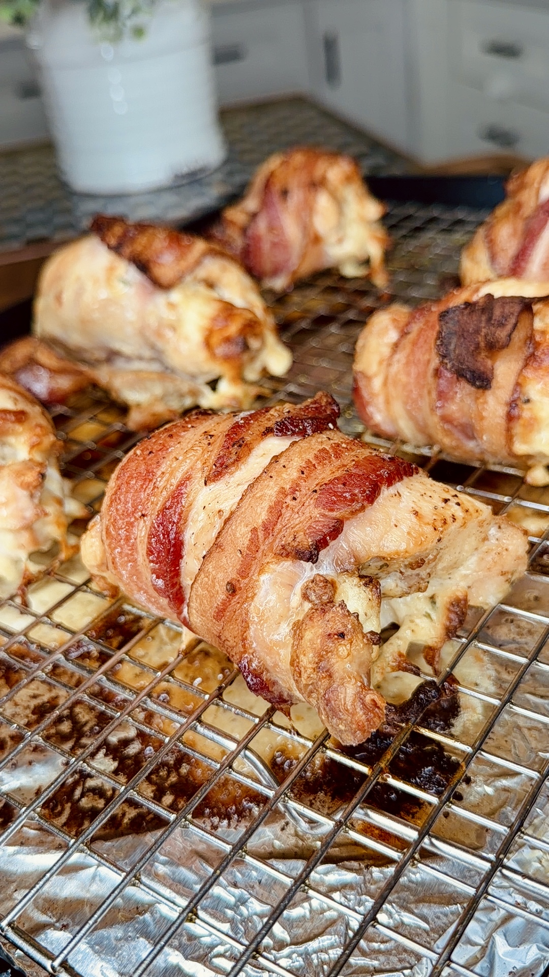 Bacon-wrapped chicken breasts are resting on a wire rack over a baking sheet, fresh out of the oven. The bacon is crispy and golden brown, with melted cheese oozing from the center of the chicken.