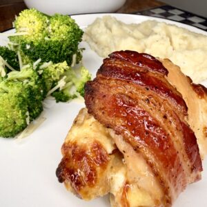 A plate of bacon-wrapped chicken breast, mashed potatoes, and steamed broccoli, with an additional bowl of broccoli in the background on a wooden table.